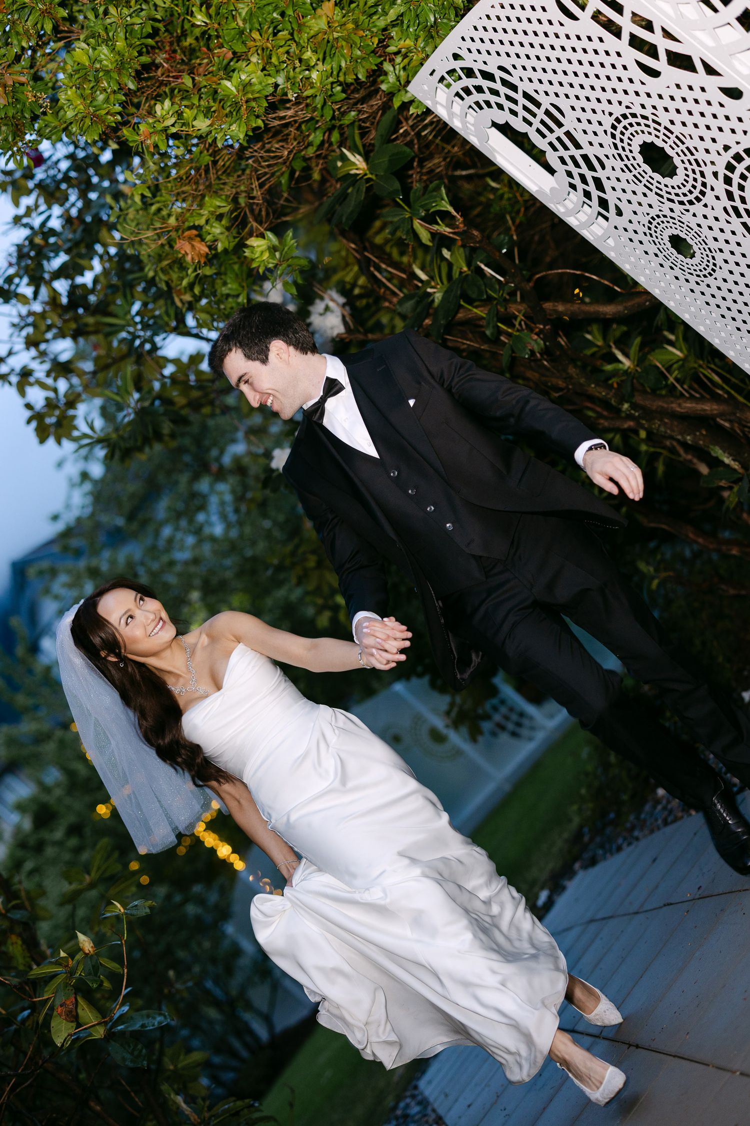 Bride in white strapless gown and groom in black tuxedo holding hands walking in garden.