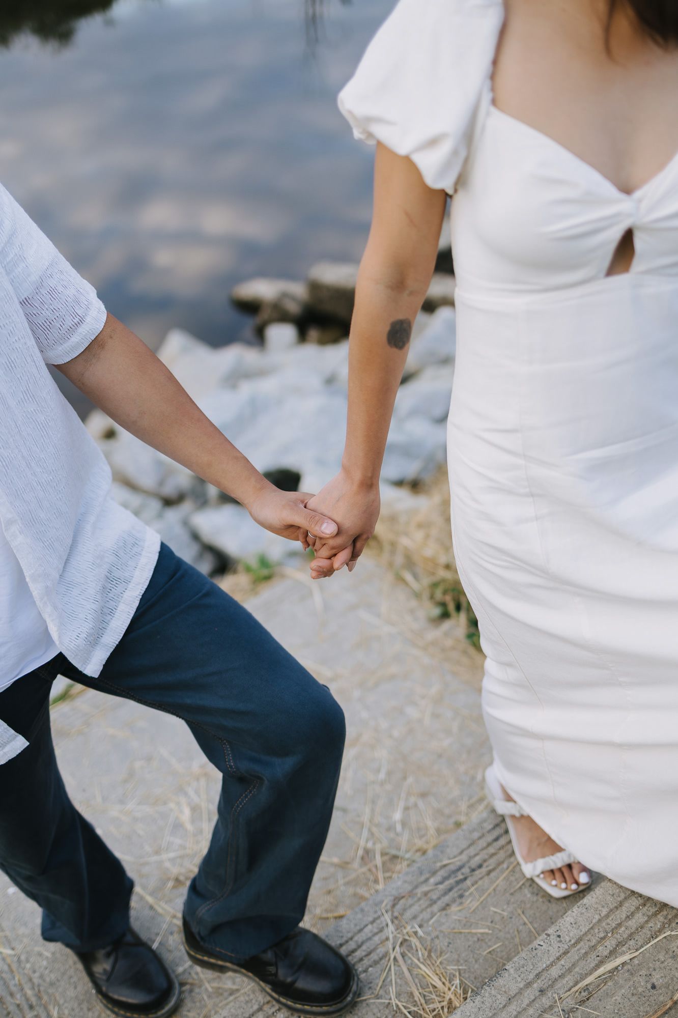 A couple in white clothing holds hands while standing on a rocky lakeside path in a romantic engagement photo.