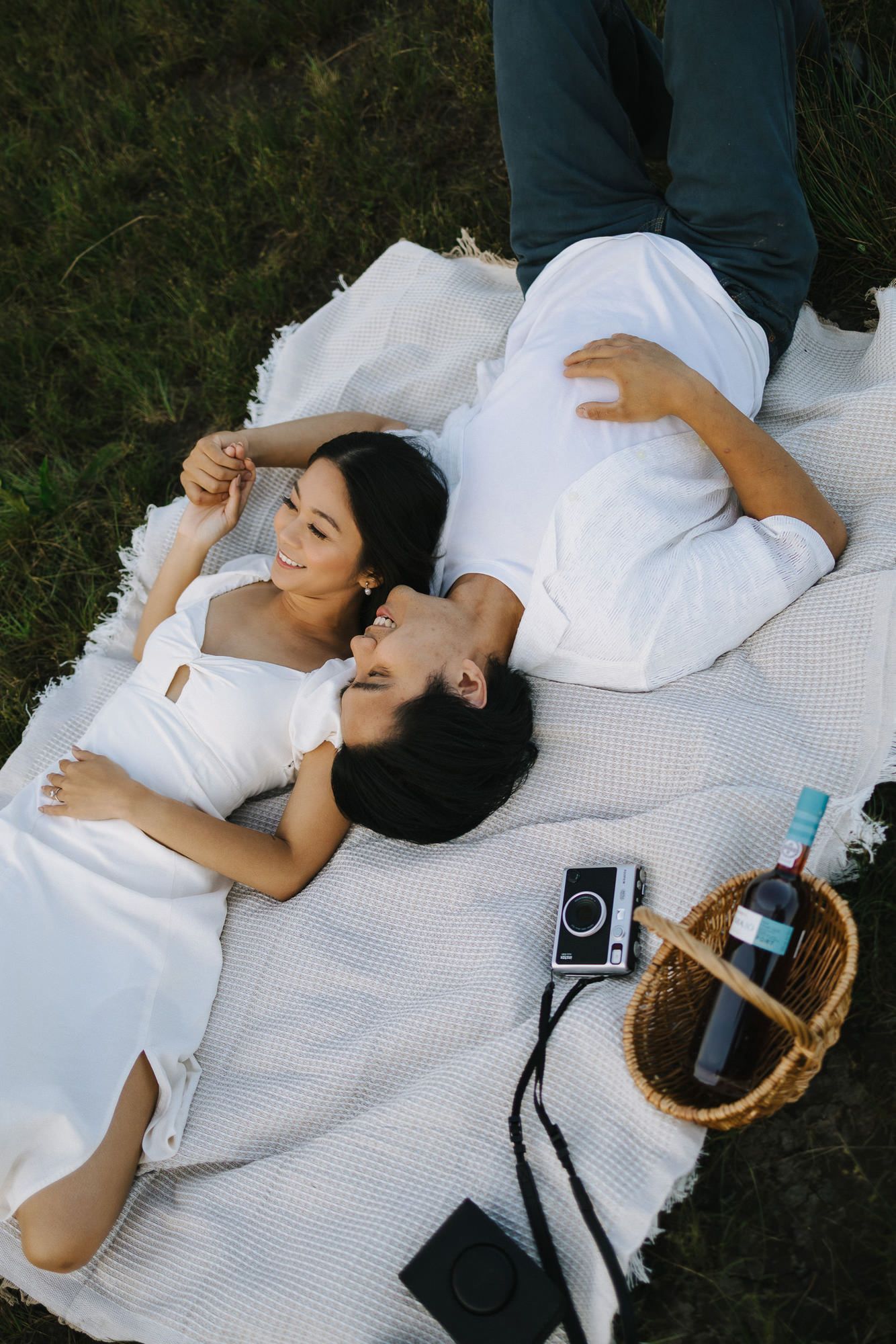 Overhead view of a smiling couple lying on a white blanket with a film camera and a picnic basket with wine.
