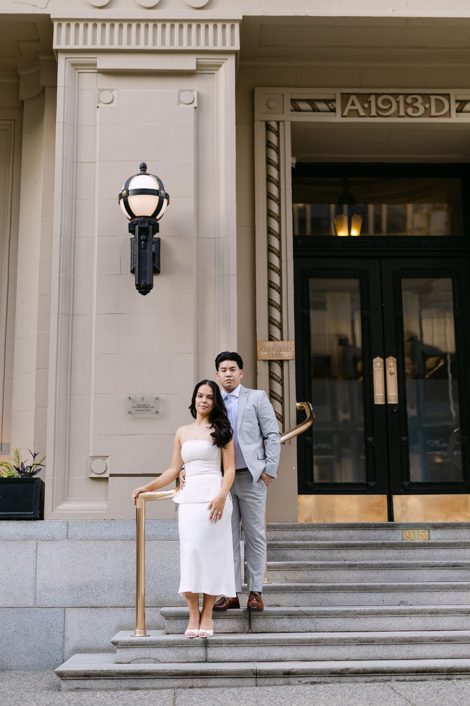 A couple in a gray suit and white dress poses on granite steps in front of an elegant building entrance marked A 1913 D.