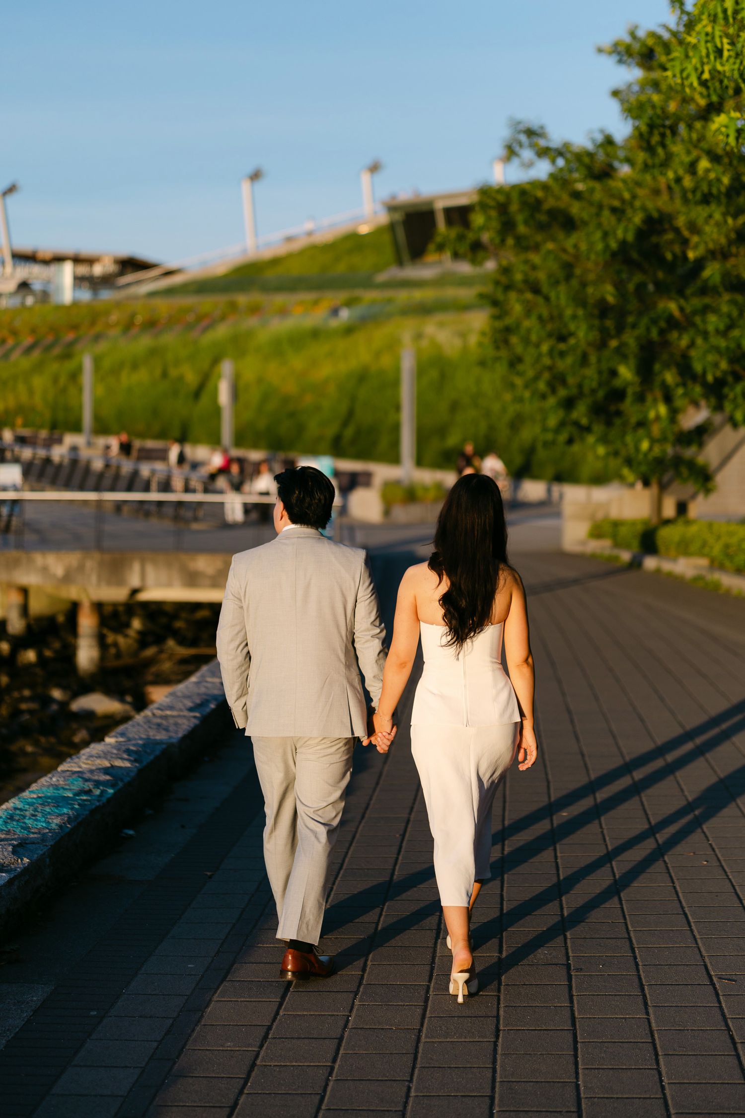 Couple holding hands walking along a sunlit waterfront promenade at golden hour, with a green grassy hillside in the background.