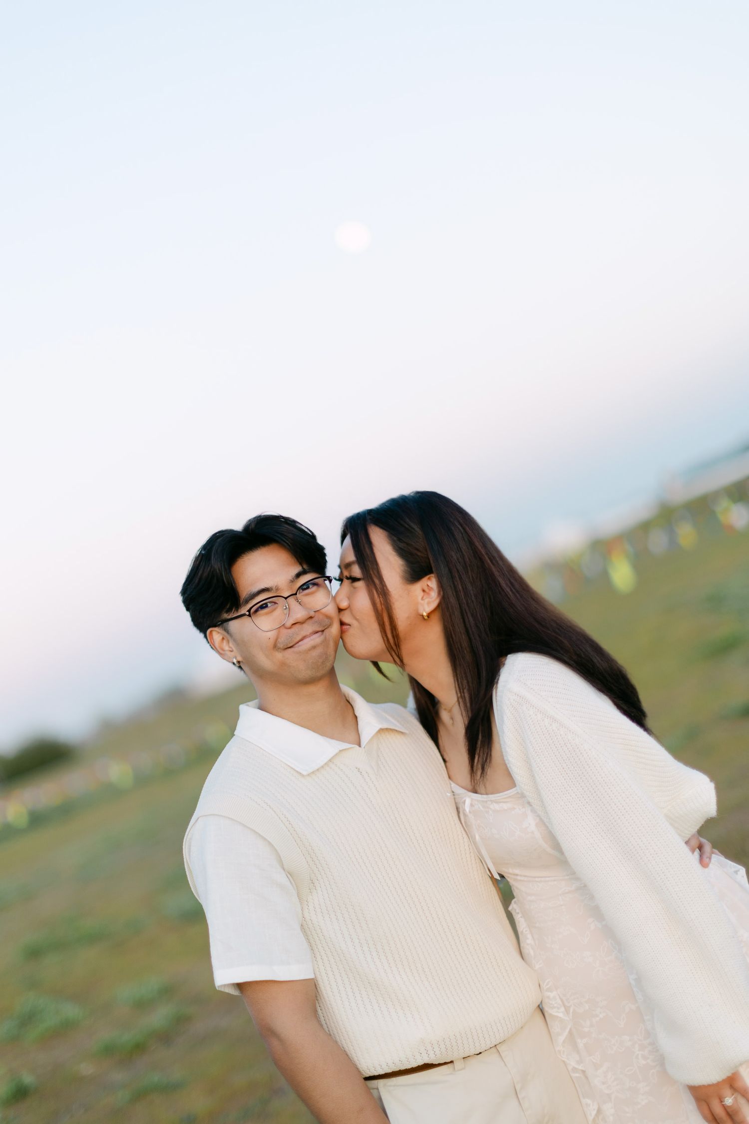 Woman in a white cardigan kisses the cheek of a smiling man in glasses and cream polo during a couples photoshoot in a grassy field at dusk.