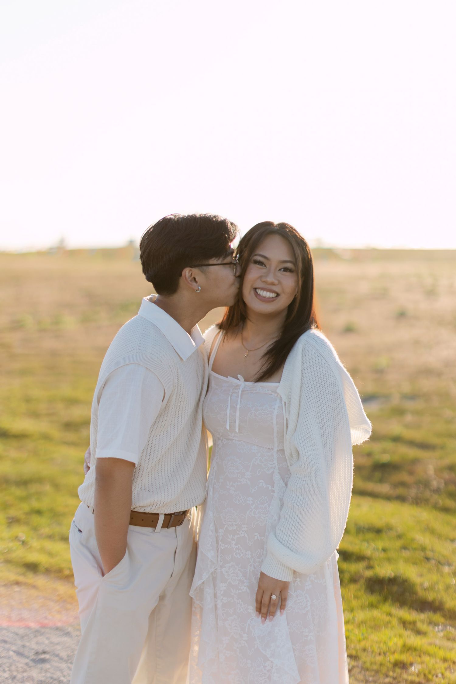 Smiling woman in a white lace dress and cardigan receives a kiss on the cheek from a partner in a cream shirt in a golden grassy field.