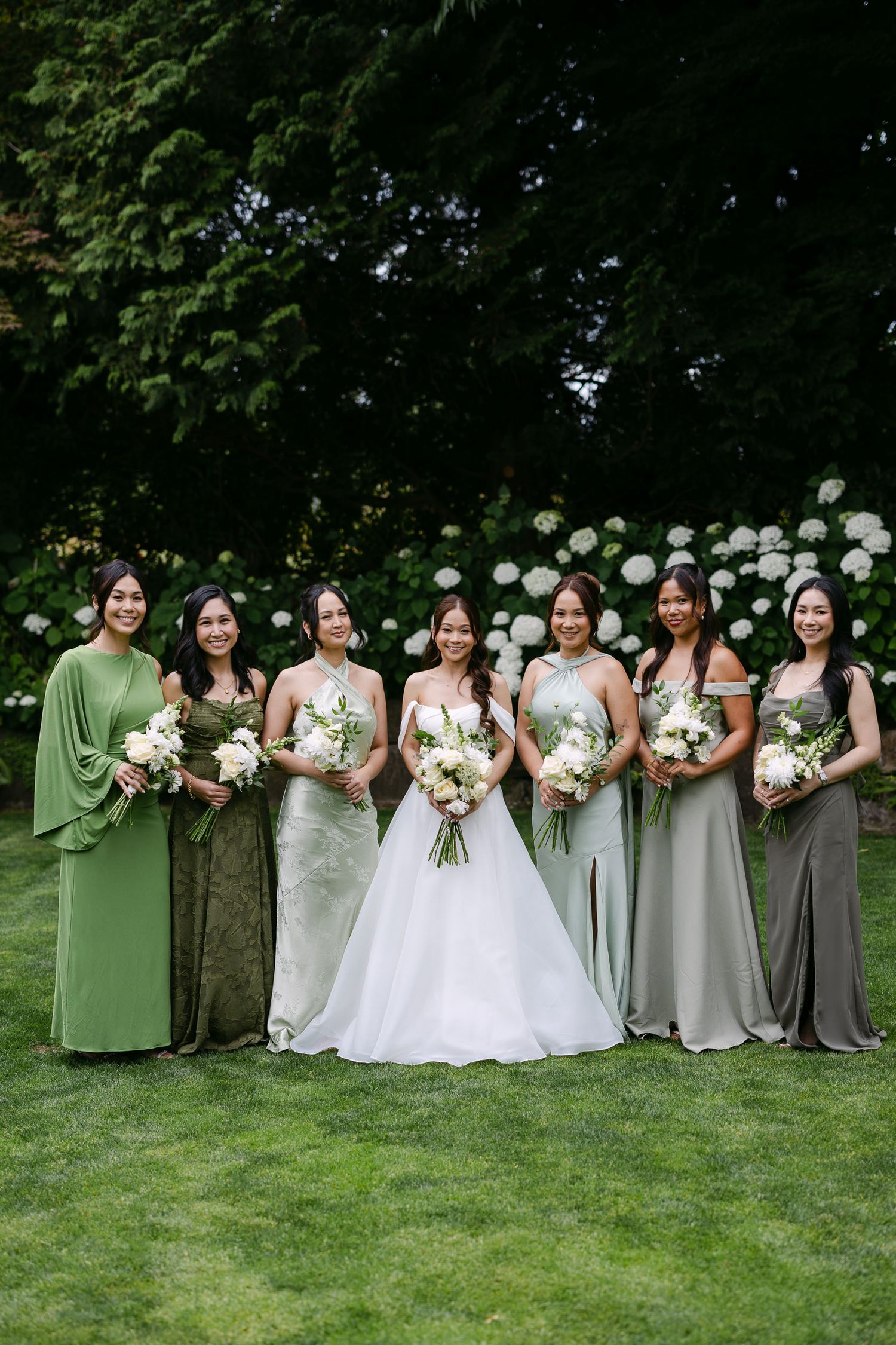 Bride in white gown with six bridesmaids in mismatched green dresses holding white bouquets outdoors.