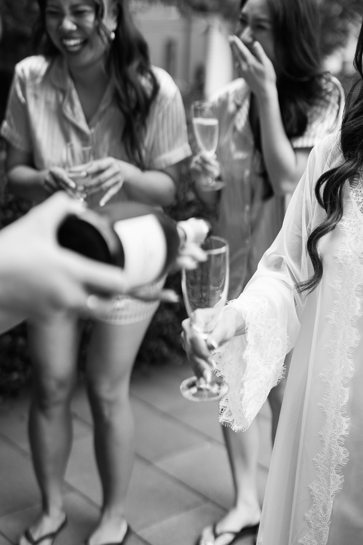 Black and white photo of bride in lace robe toasting champagne with bridesmaids at a wedding celebration.