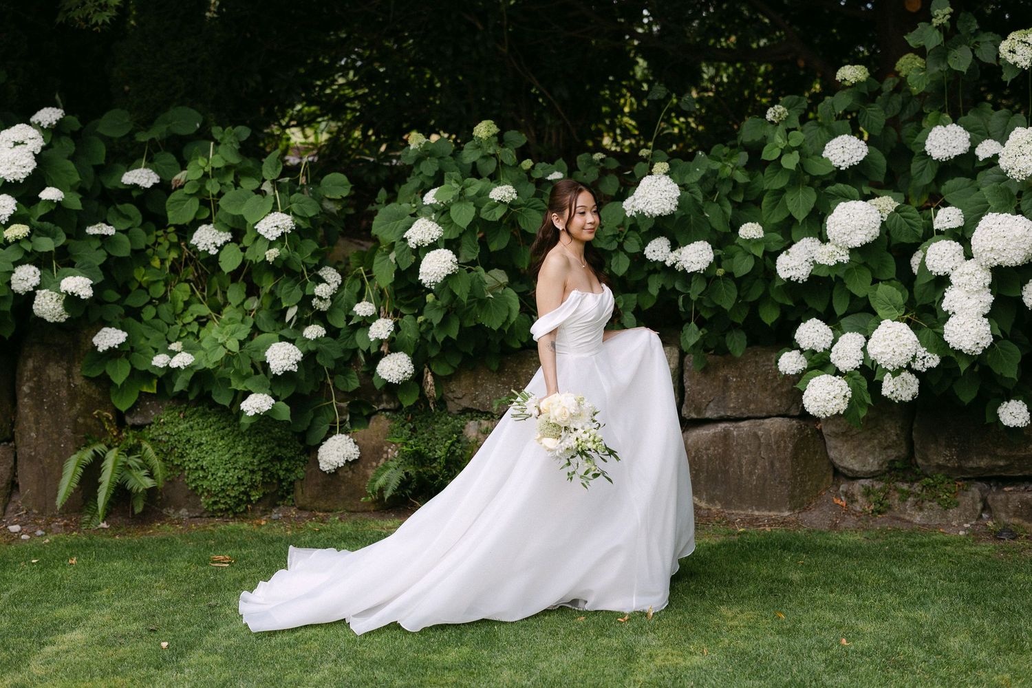 Bride in an off-shoulder white gown standing before a lush hydrangea garden wall holding a bouquet.