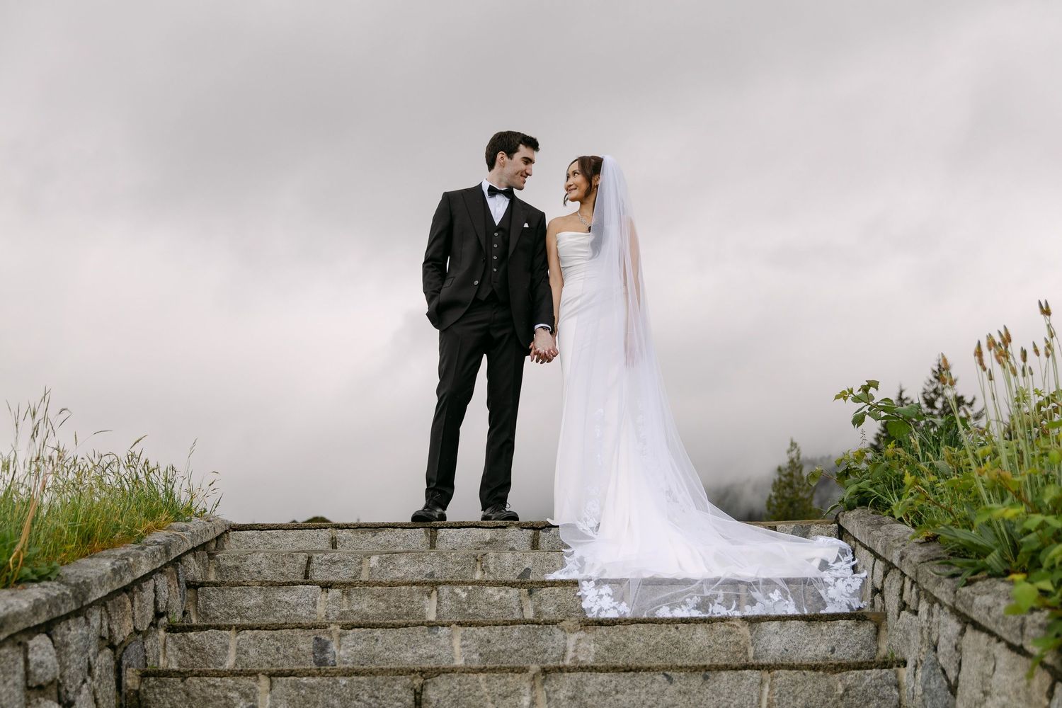 Bride and groom stand on stone steps holding hands with misty sky backdrop on their wedding day.