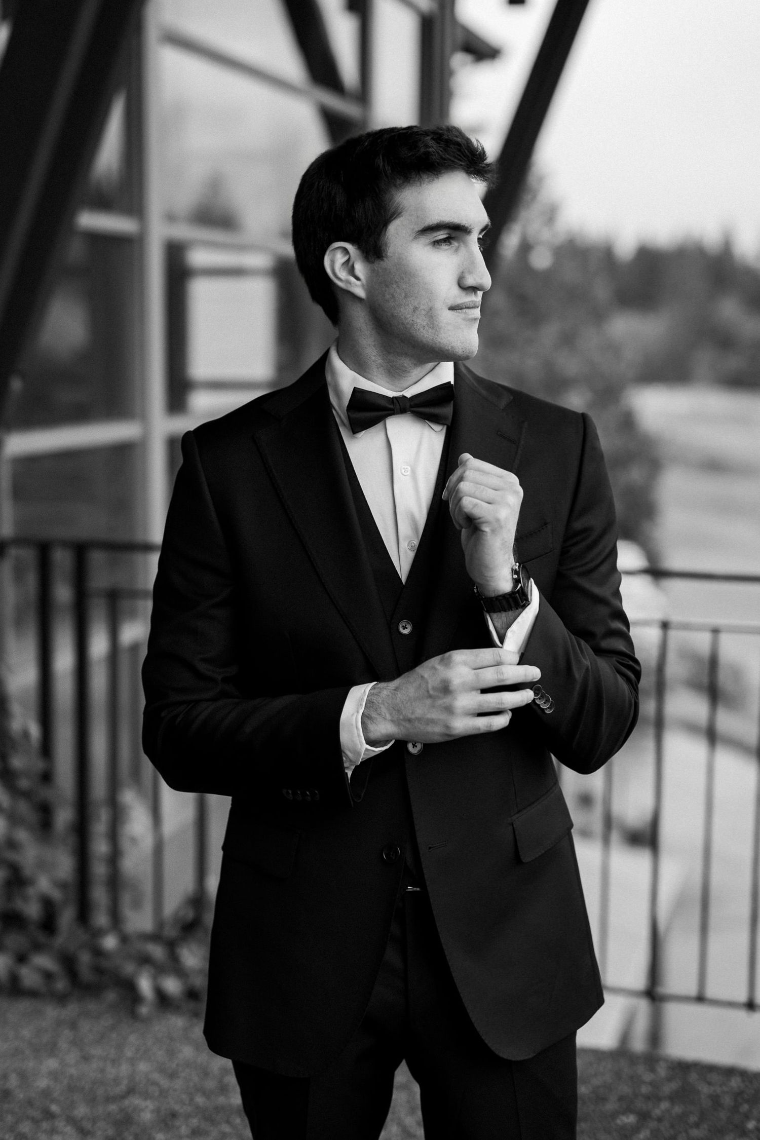 Young man in black tuxedo and bow tie adjusting cufflinks outdoors in elegant black and white portrait photo.