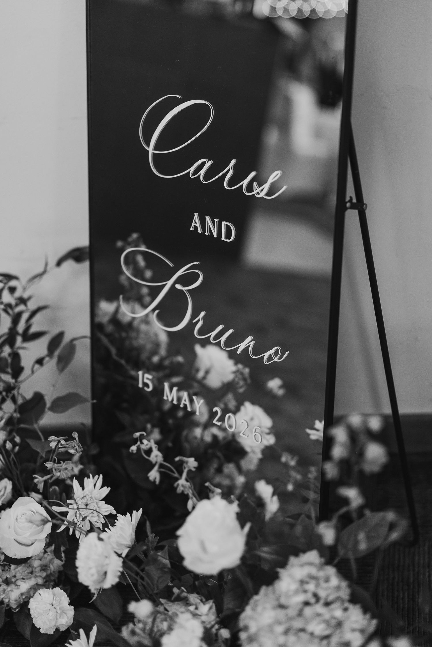 Black and white wedding welcome sign for Caro and Bruno, dated 15 May, surrounded by elegant floral arrangements.