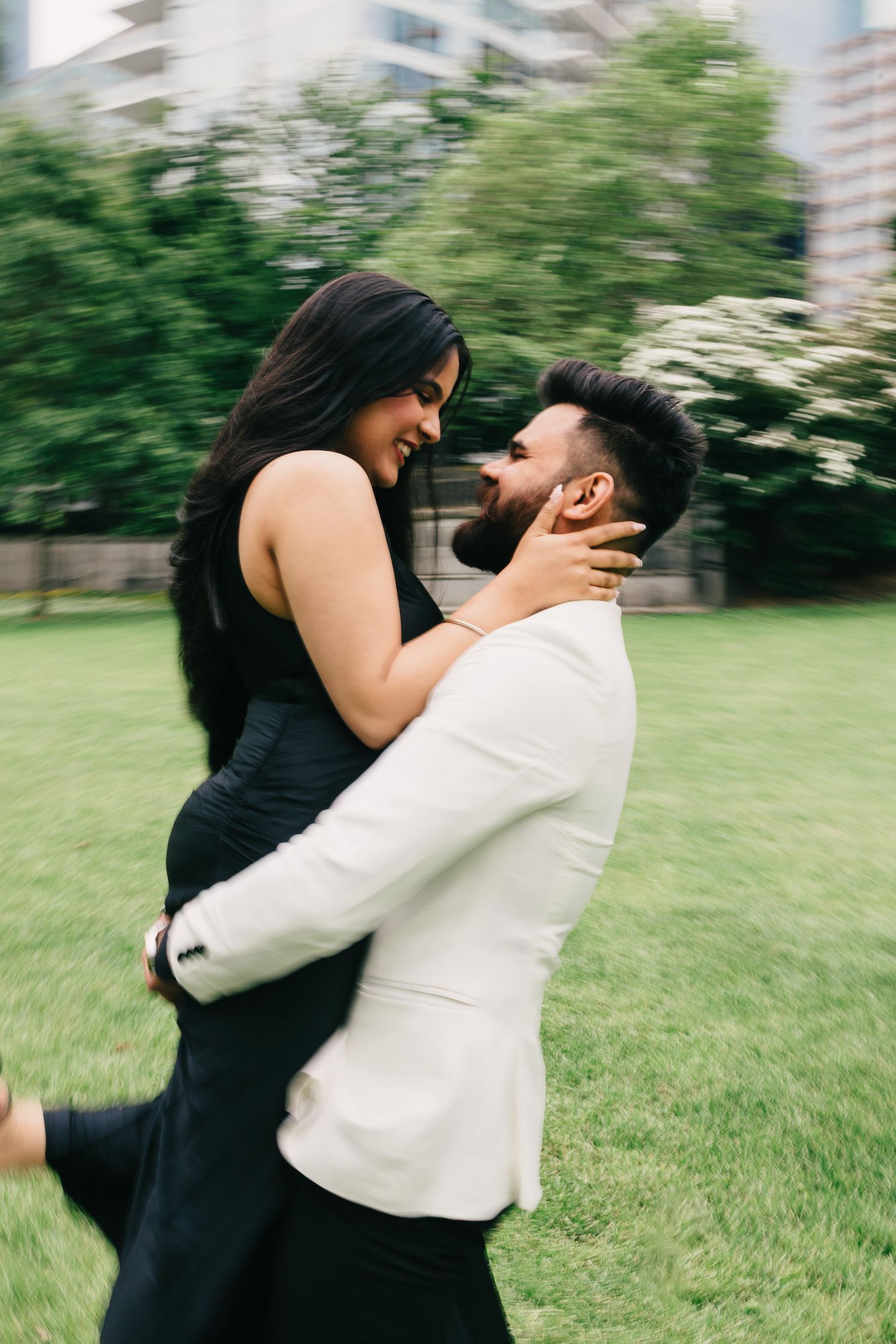 A couple in formal attire embraces joyfully in a green outdoor setting, spinning in celebration.