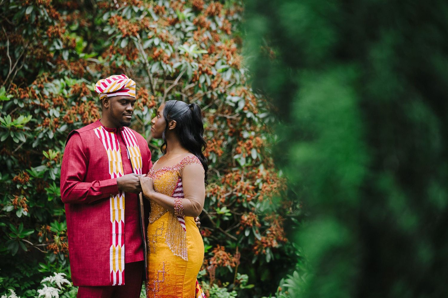 A couple in traditional formal attire share an intimate moment in a lush garden setting with orange flowers.