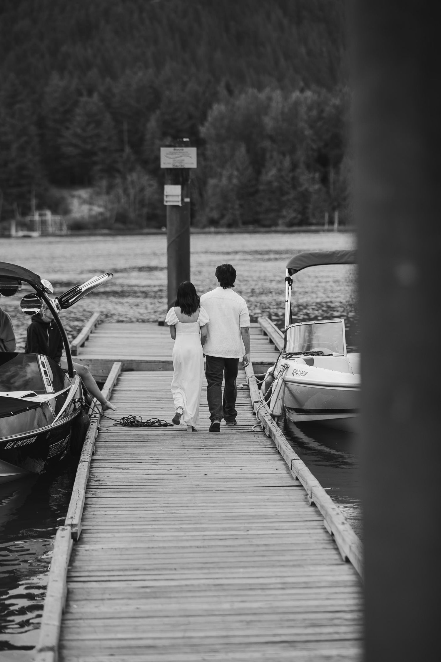 Black and white photo of a person walking on a wooden dock between moored boats at a marina.