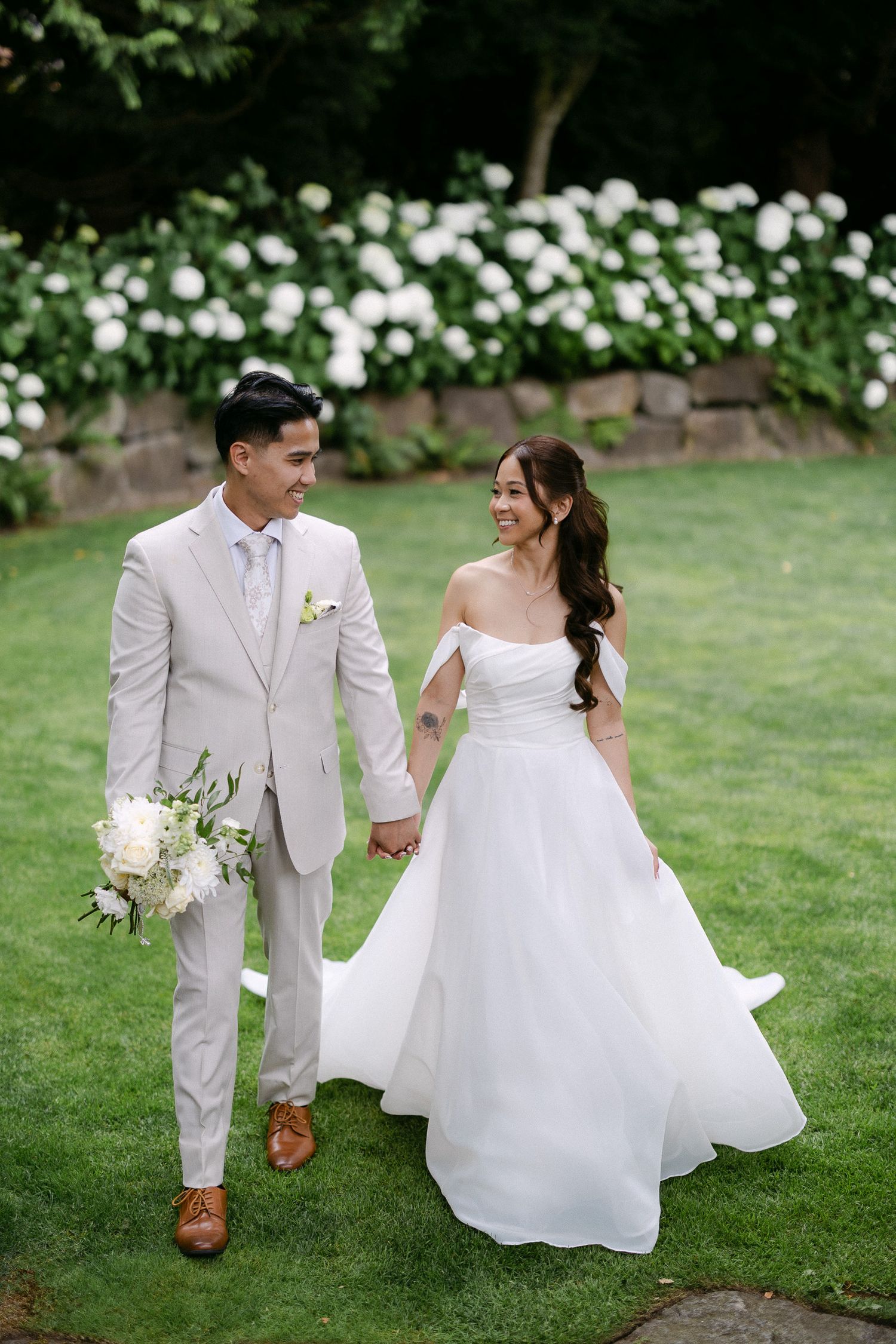 Couple walks hand in hand on green lawn, groom in beige suit, bride in white off-shoulder gown, white hydrangeas behind.