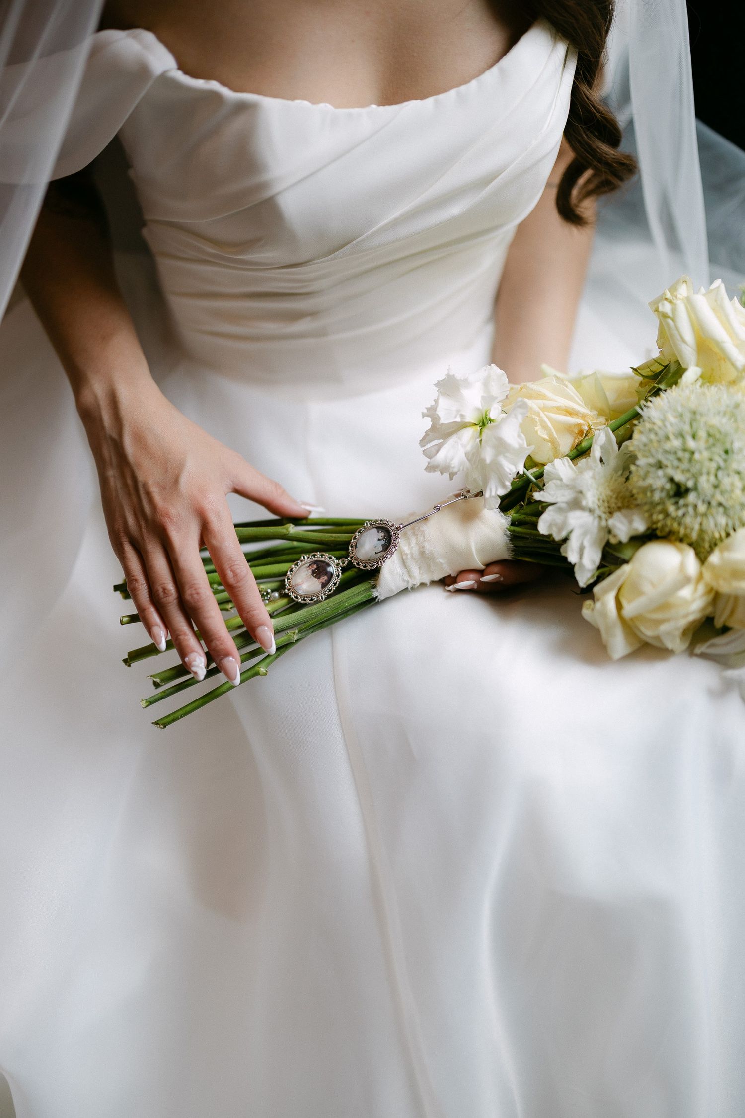 Bride in white off-shoulder gown holding elegant bouquet of white roses and greenery with decorative brooch detail.