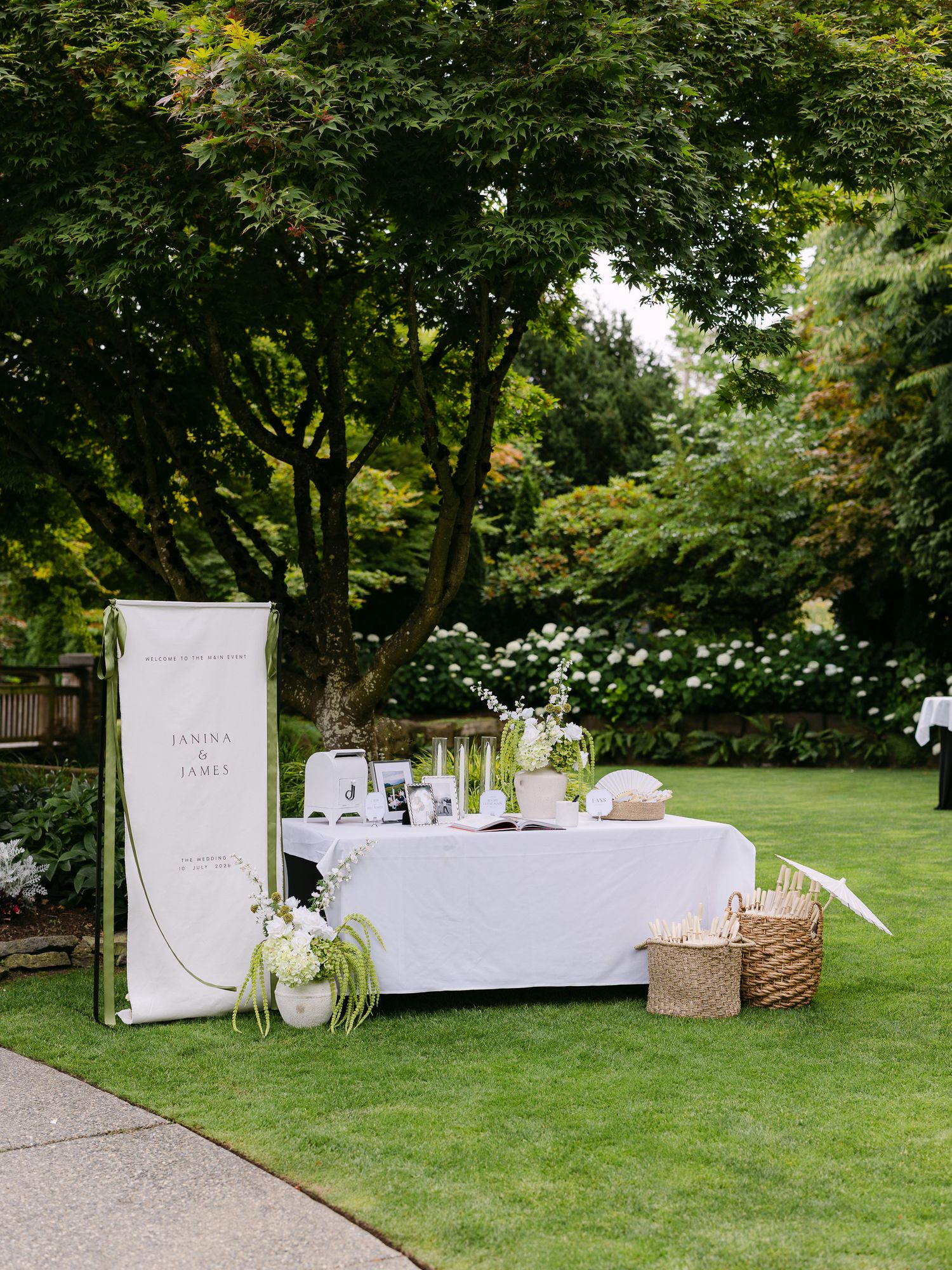 Elegant outdoor wedding welcome table with white linen, floral arrangements, and a personalized banner on a lush green lawn.