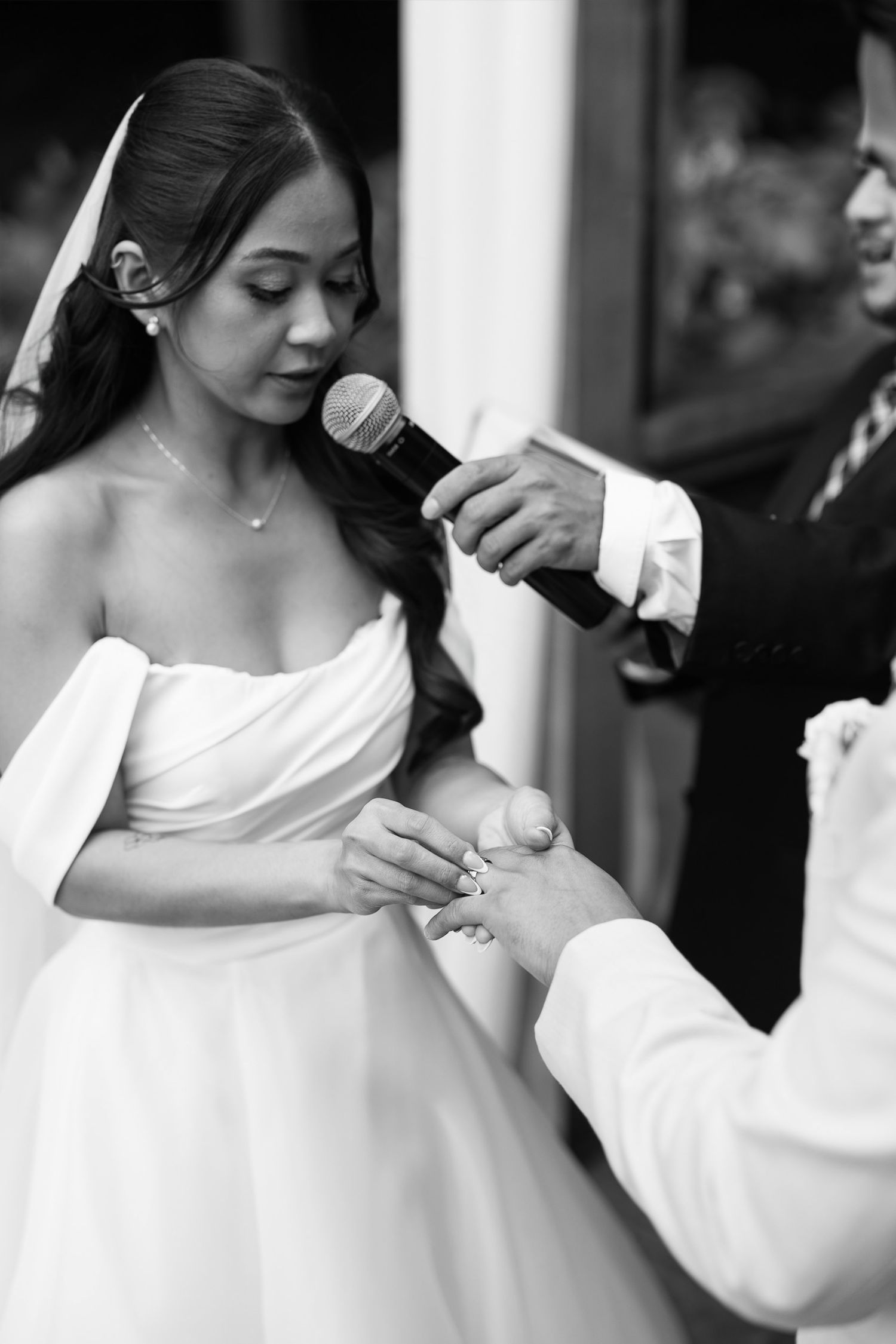 Bride places wedding ring on groom's finger during emotional outdoor ceremony in black and white.