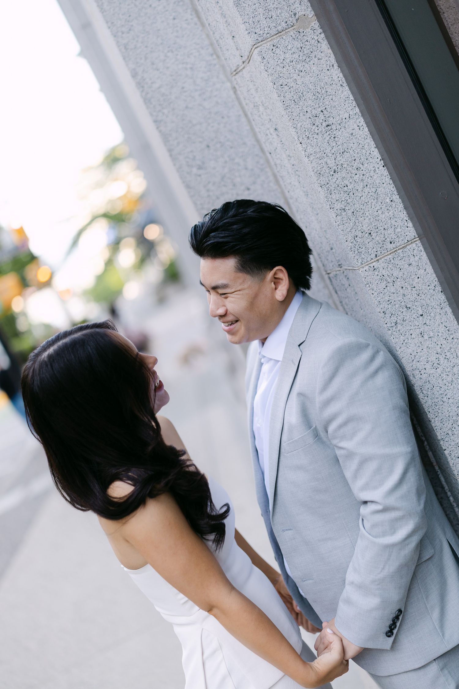 Laughing couple holding hands lean against a stone wall on a city street, captured from an angle in warm natural light.