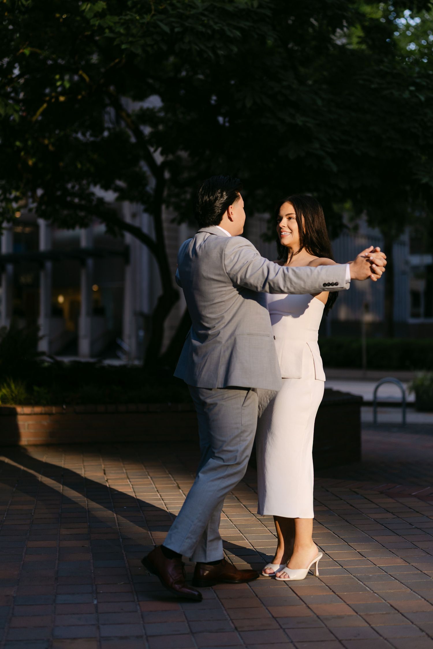 Couple in a gray suit and white dress dance together on a sunlit brick plaza beneath trees during an engagement photo session.