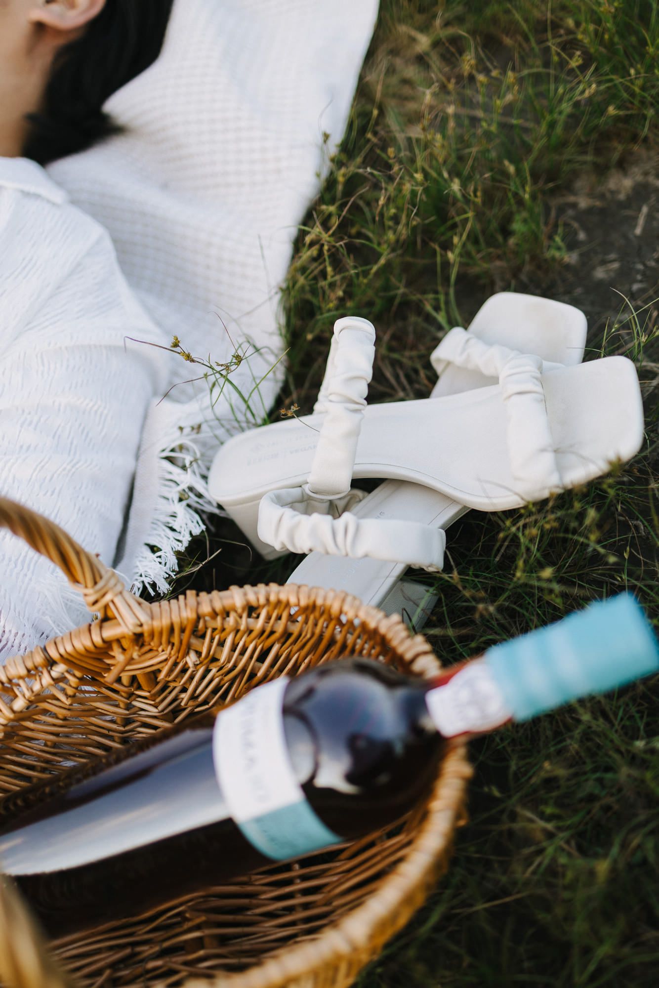 Picnic scene with white sandals, a wine bottle in a wicker basket, and a woman resting on a blanket in the grass.