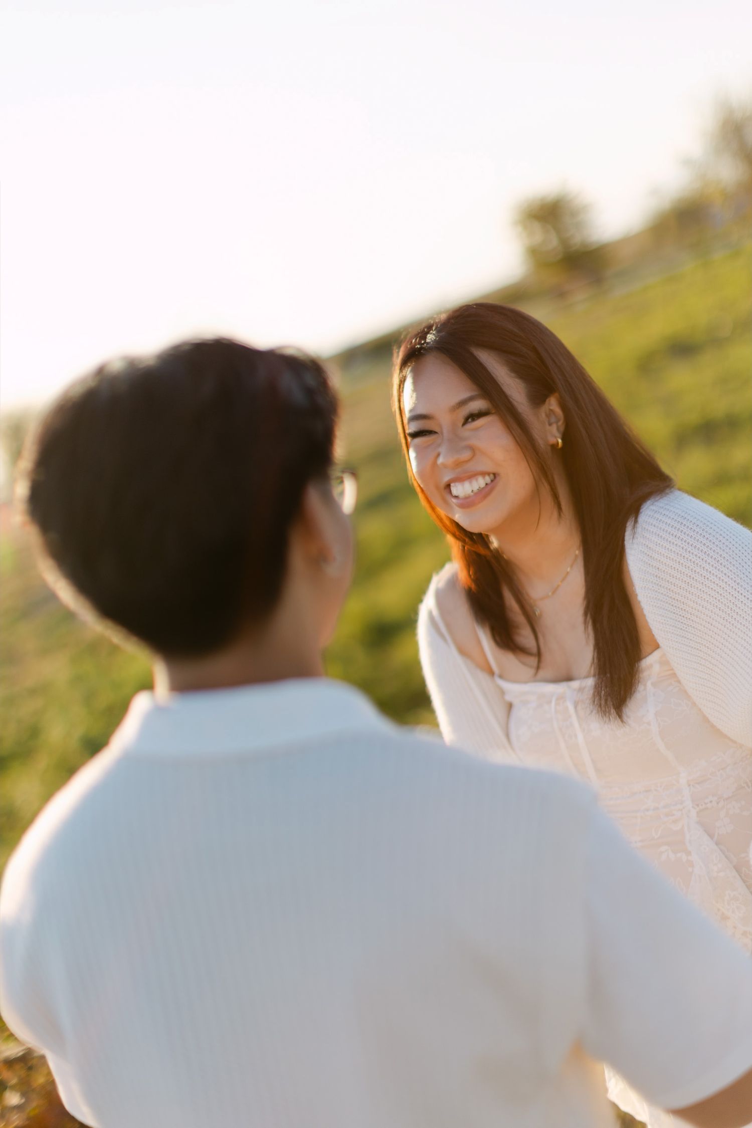 Smiling woman with long brown hair in a white lace top laughs with a person in white in a golden sunlit meadow.