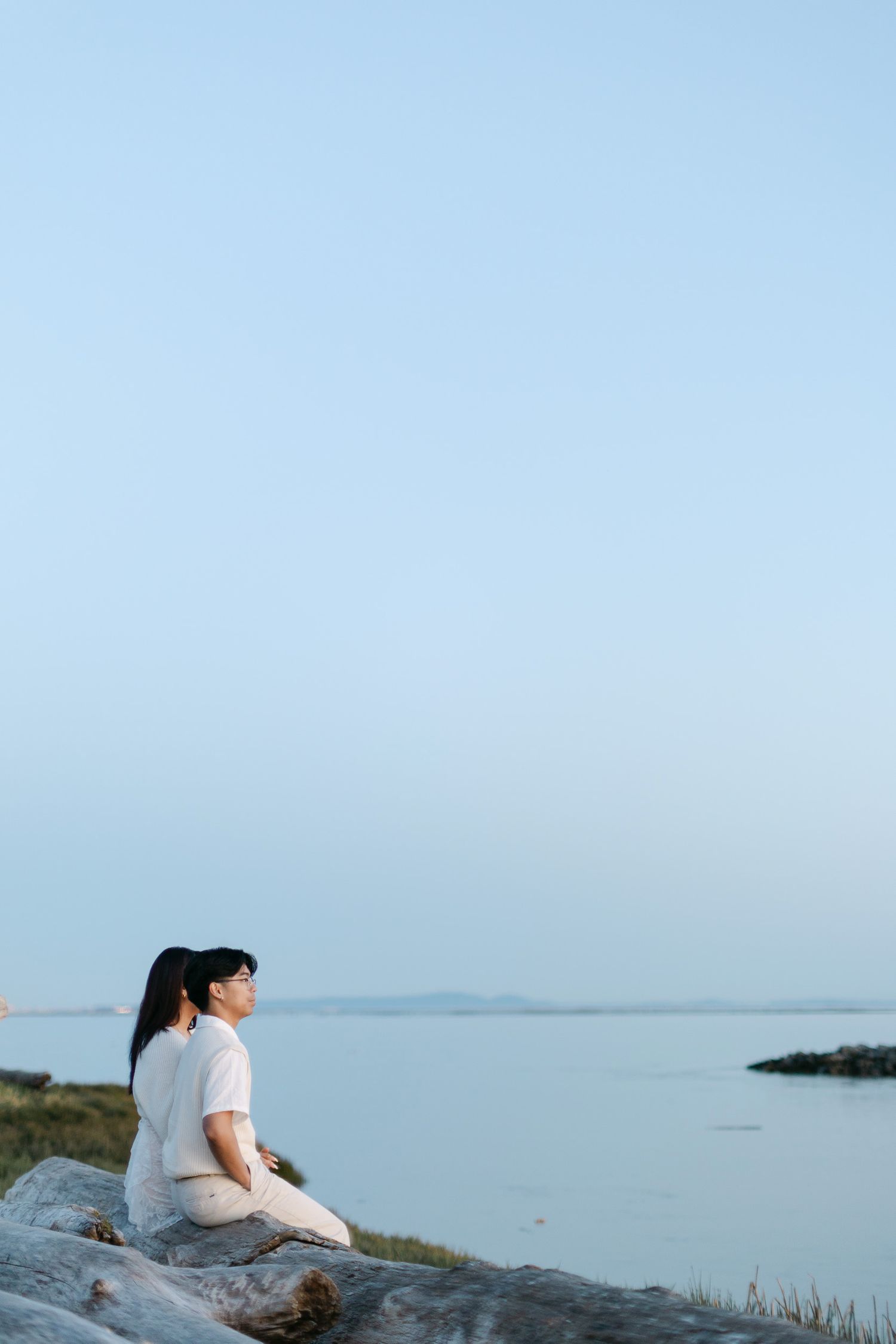 Two people in light clothing sit close together on a driftwood log, gazing across calm water under a soft, pale blue sky at dusk.
