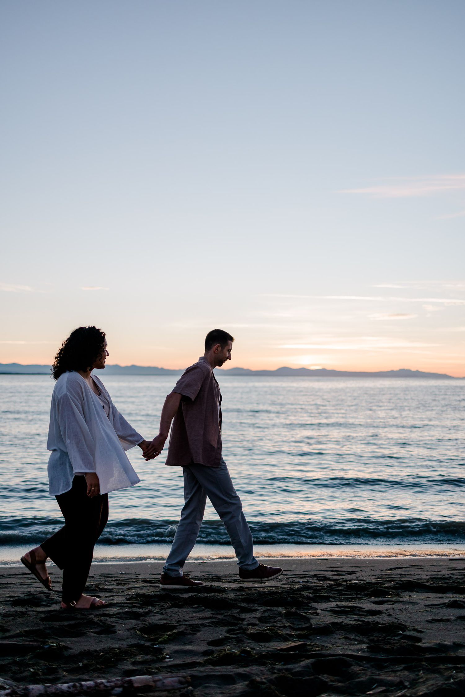 Two silhouettes sit on a dark beach at sunset with ocean waves and mountains in the background.