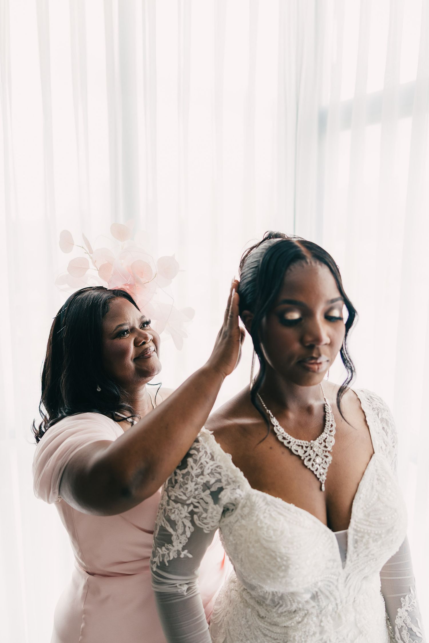 Two women share an intimate moment while getting ready, one wearing a pink dress and one in a white wedding gown.