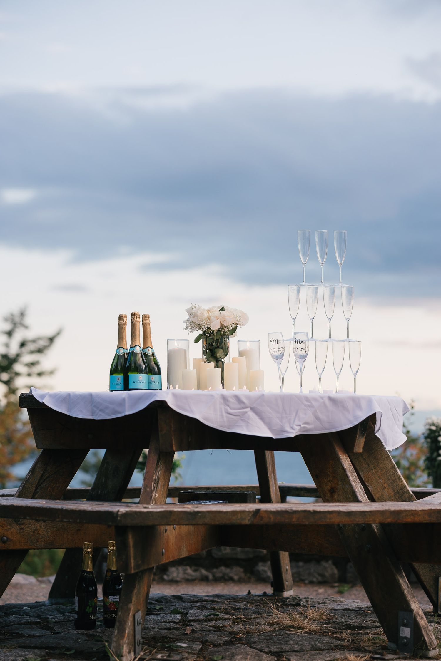 Rustic wooden tables set up outdoors with champagne bottles, glasses, and white candles against a cloudy sky.