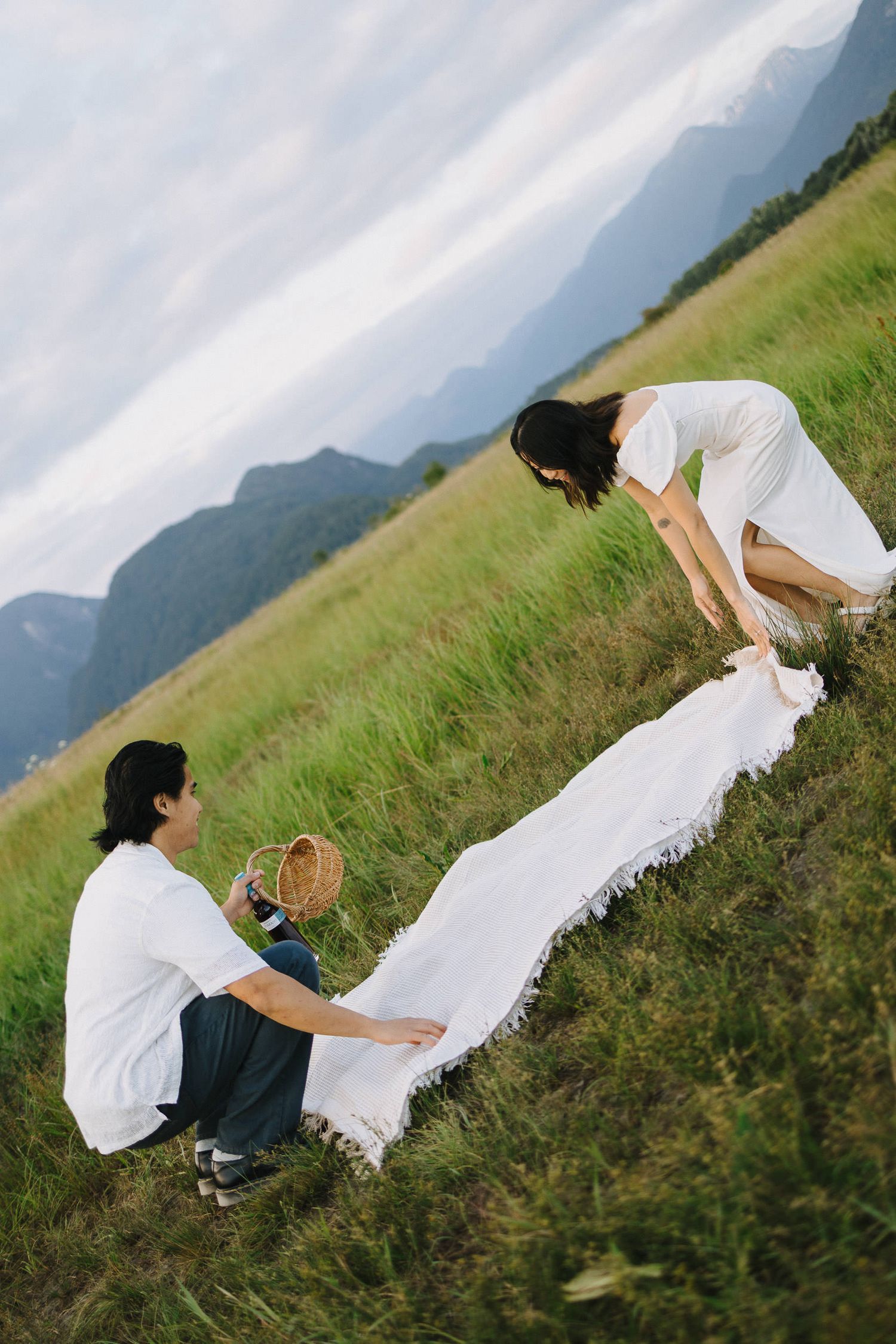 A couple in white outfits playfully poses on a grassy hillside with mountains in the background.