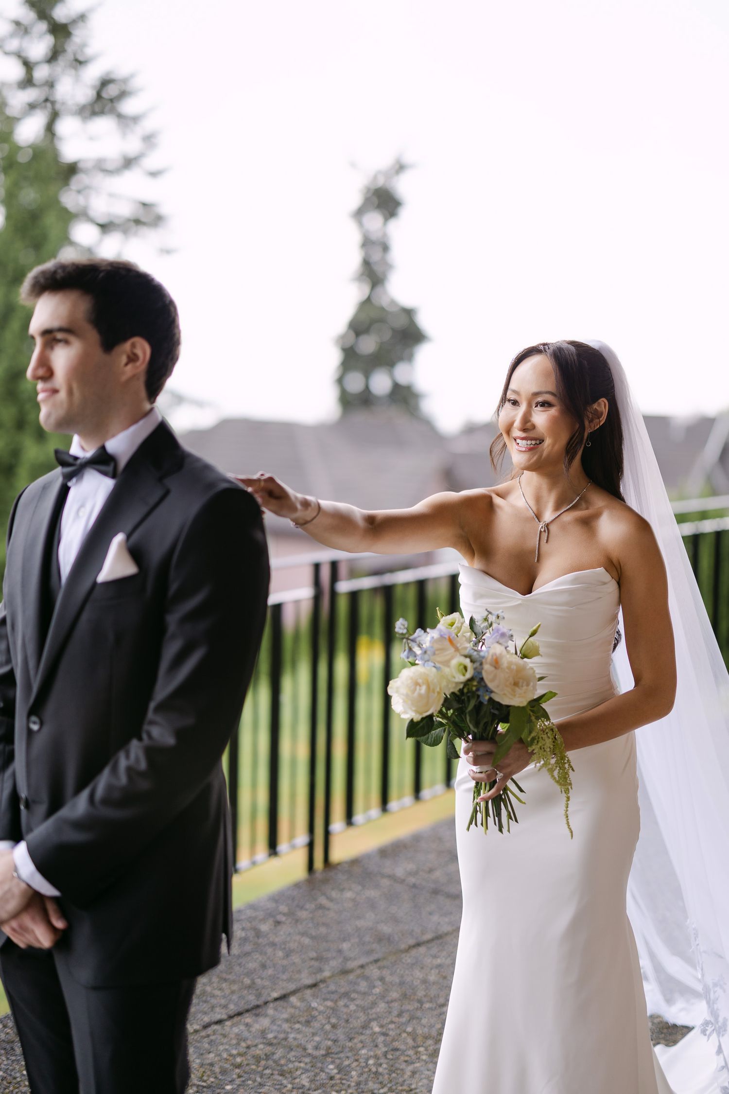 Bride in white strapless gown holds bouquet, tapping groom's shoulder during first look on outdoor terrace.