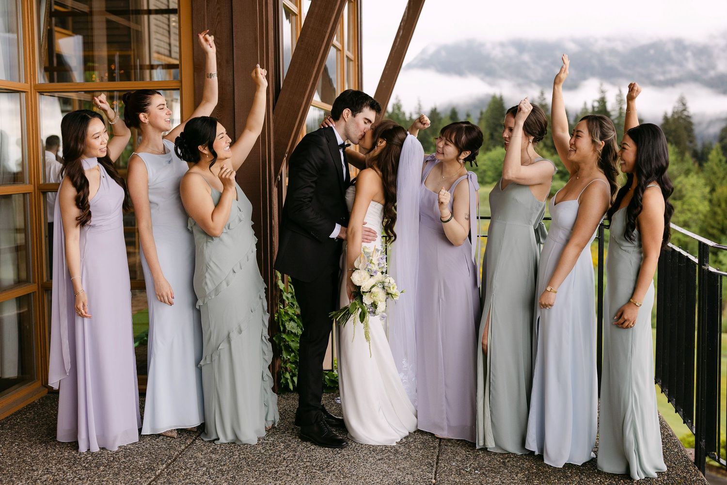 Bride and groom kiss surrounded by cheering bridesmaids in lavender and sage dresses at an elegant mountain wedding venue.