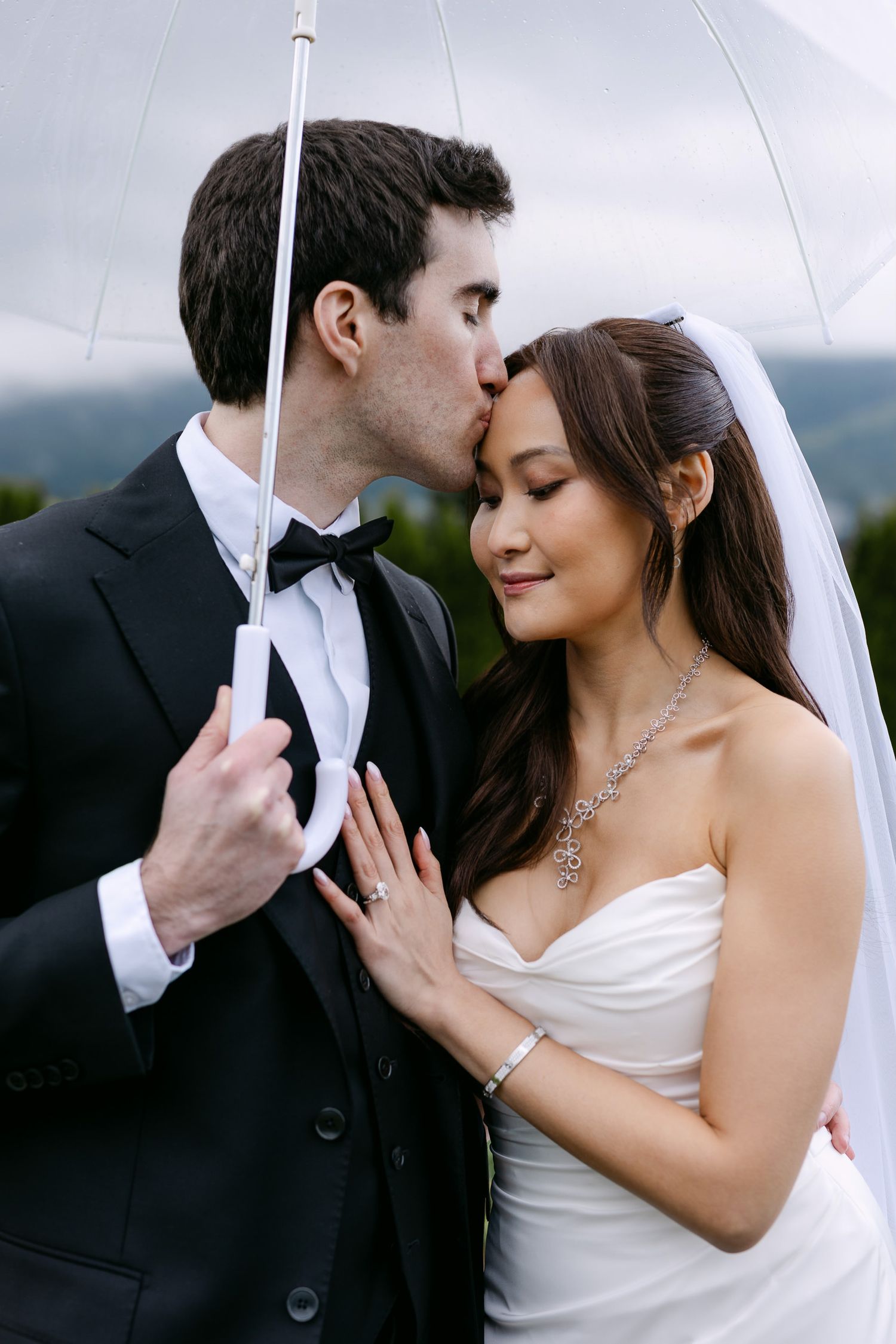 Bride and groom share a romantic kiss under a clear umbrella on their rainy wedding day.