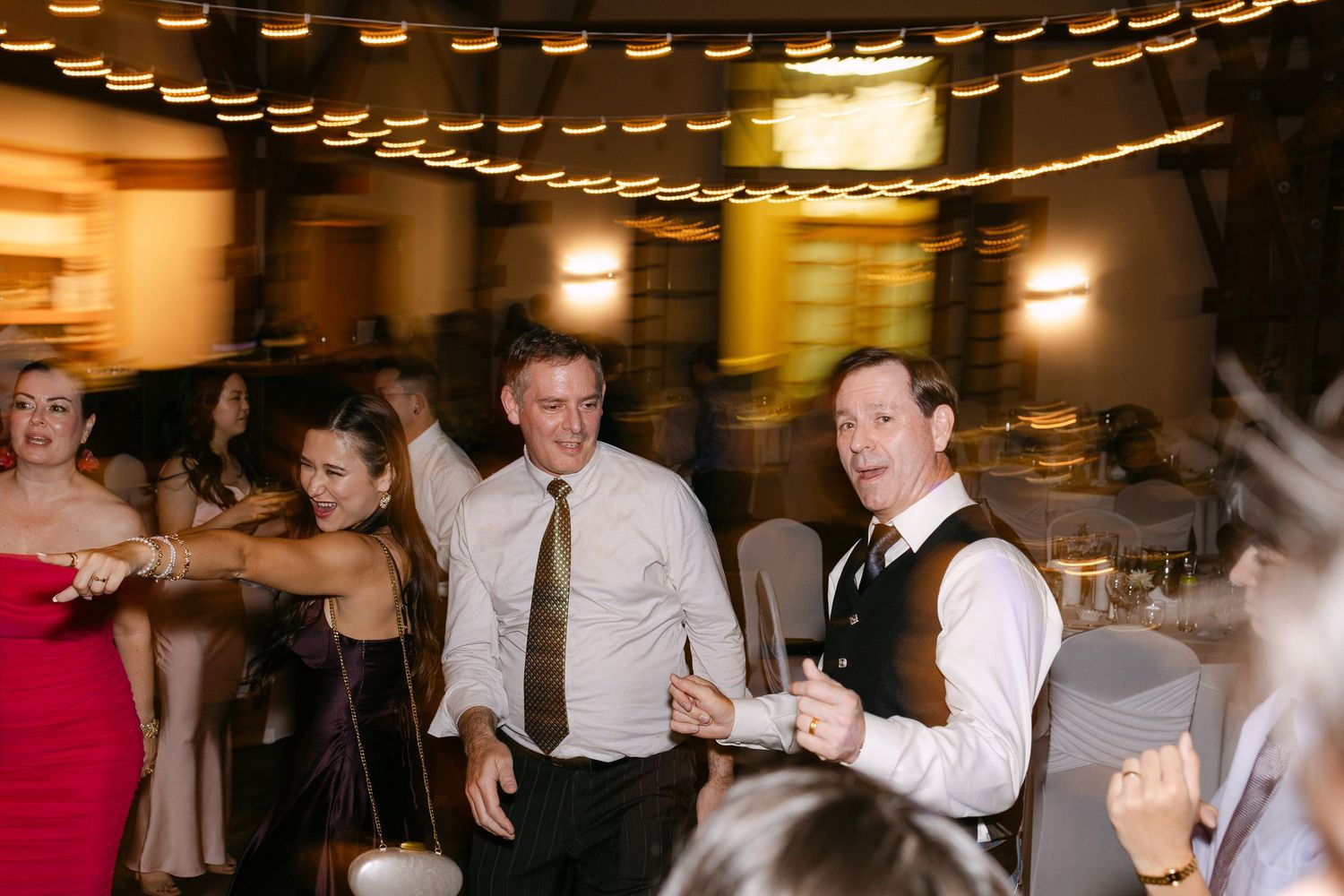 Guests dancing and celebrating joyfully at a wedding reception in a warmly lit venue.