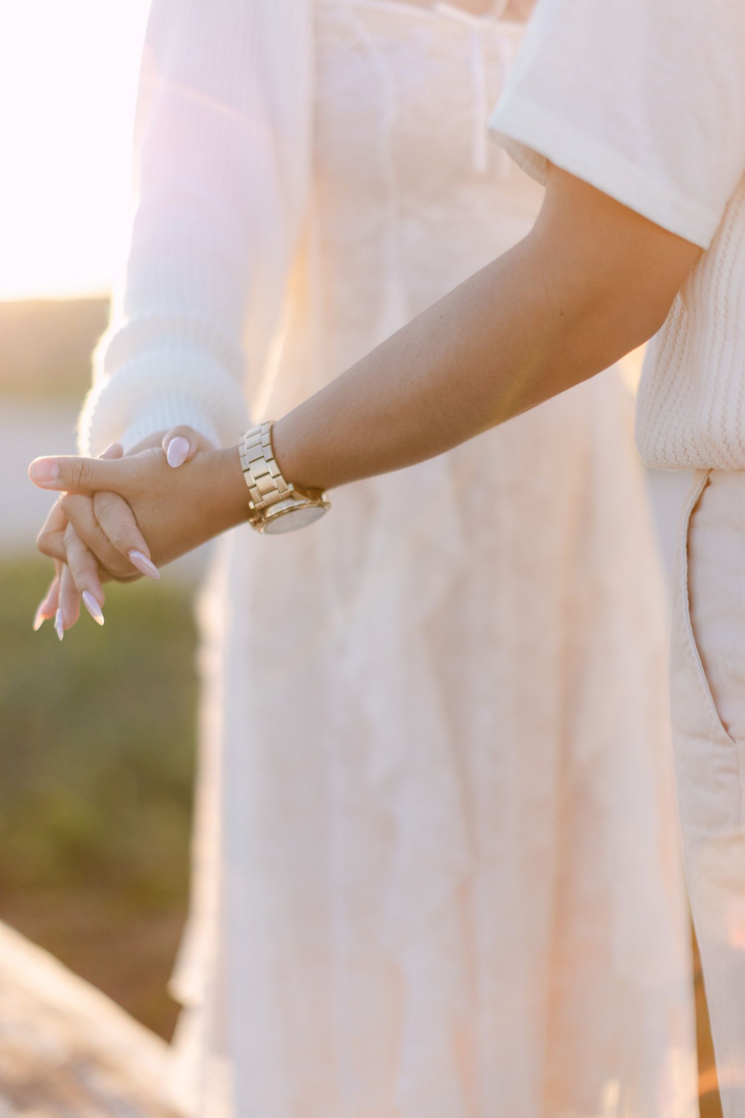Close-up of a couple holding hands at sunset, with manicured nails and a gold watch, in soft cream outfits.