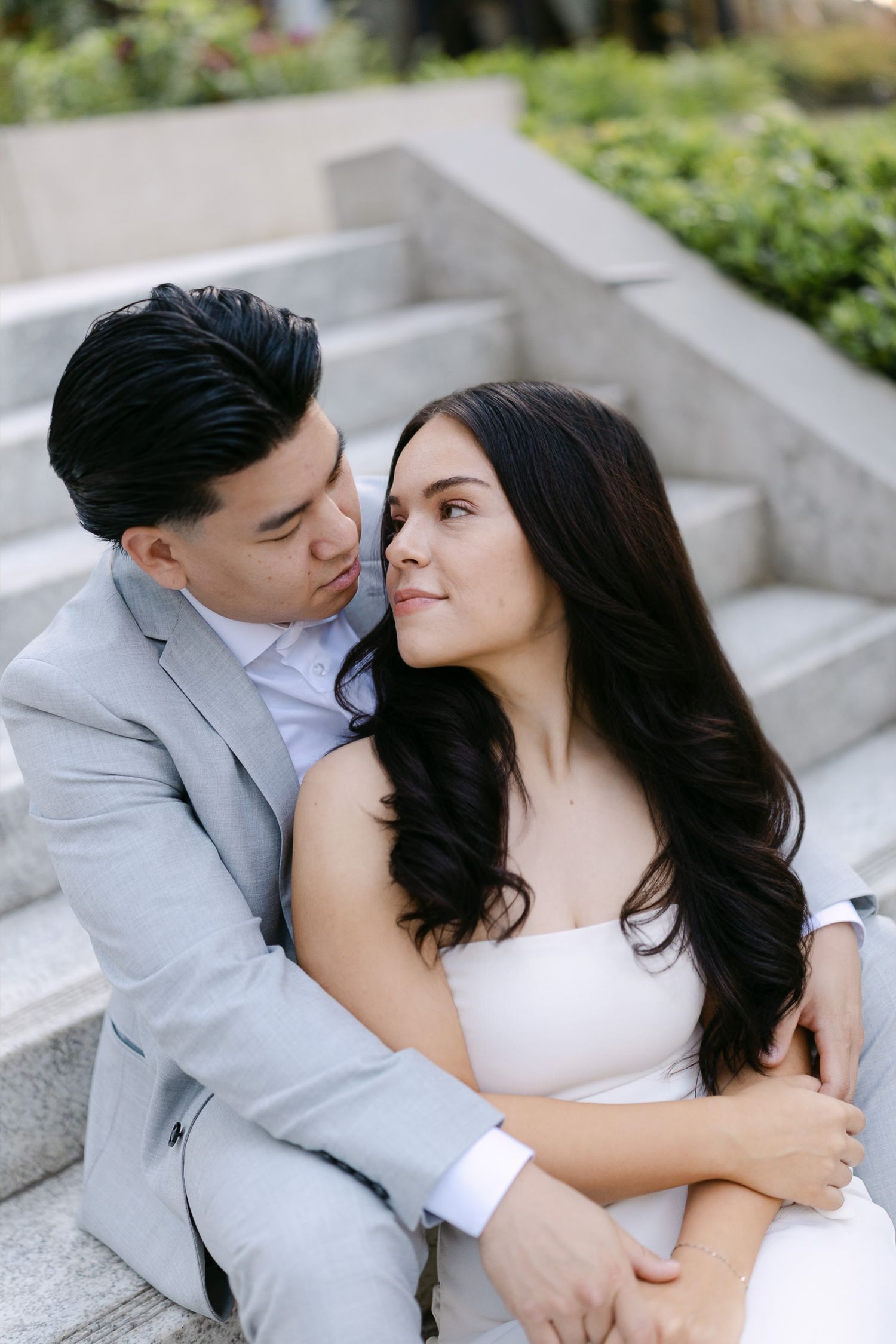 Couple in light gray suit and white dress sit close on outdoor stone steps, sharing a tender moment during an engagement photo session.