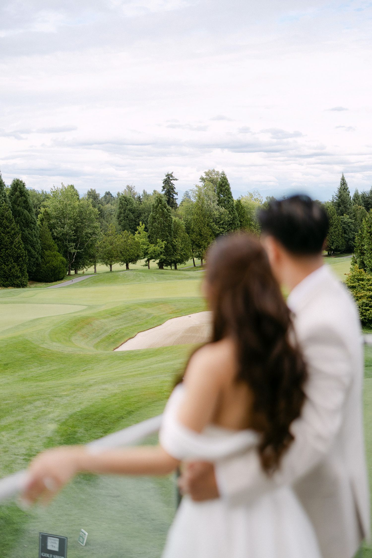 Couple in wedding attire embracing on a balcony overlooking a lush green golf course with tall trees.