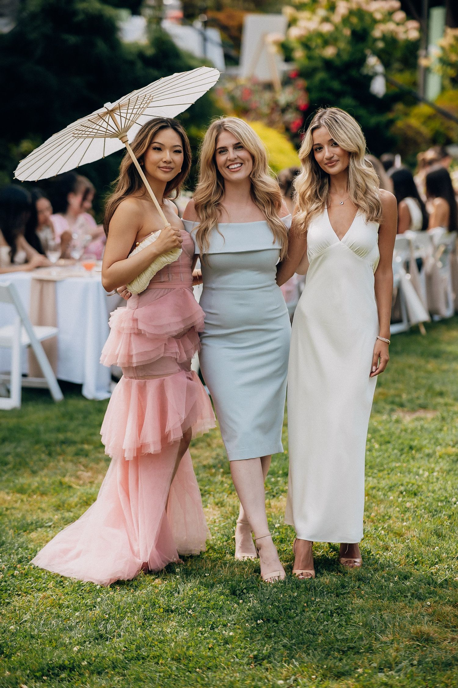 A group of friends in pastel dresses pose together on a grassy lawn holding white parasols at an outdoor event.