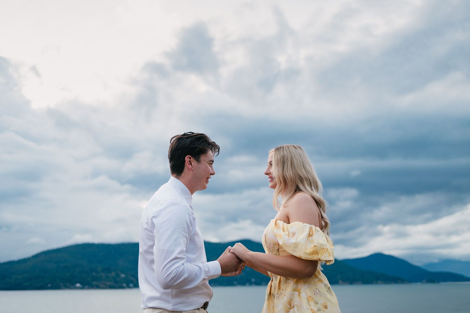 A couple shares a romantic moment by a lake with mountains and dramatic cloudy sky in the background.