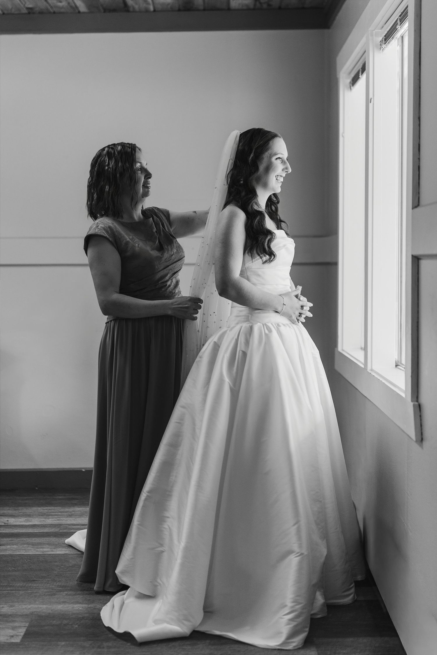 Black and white photo of a bride in a white gown and veil being helped by a bridesmaid near a bright window.