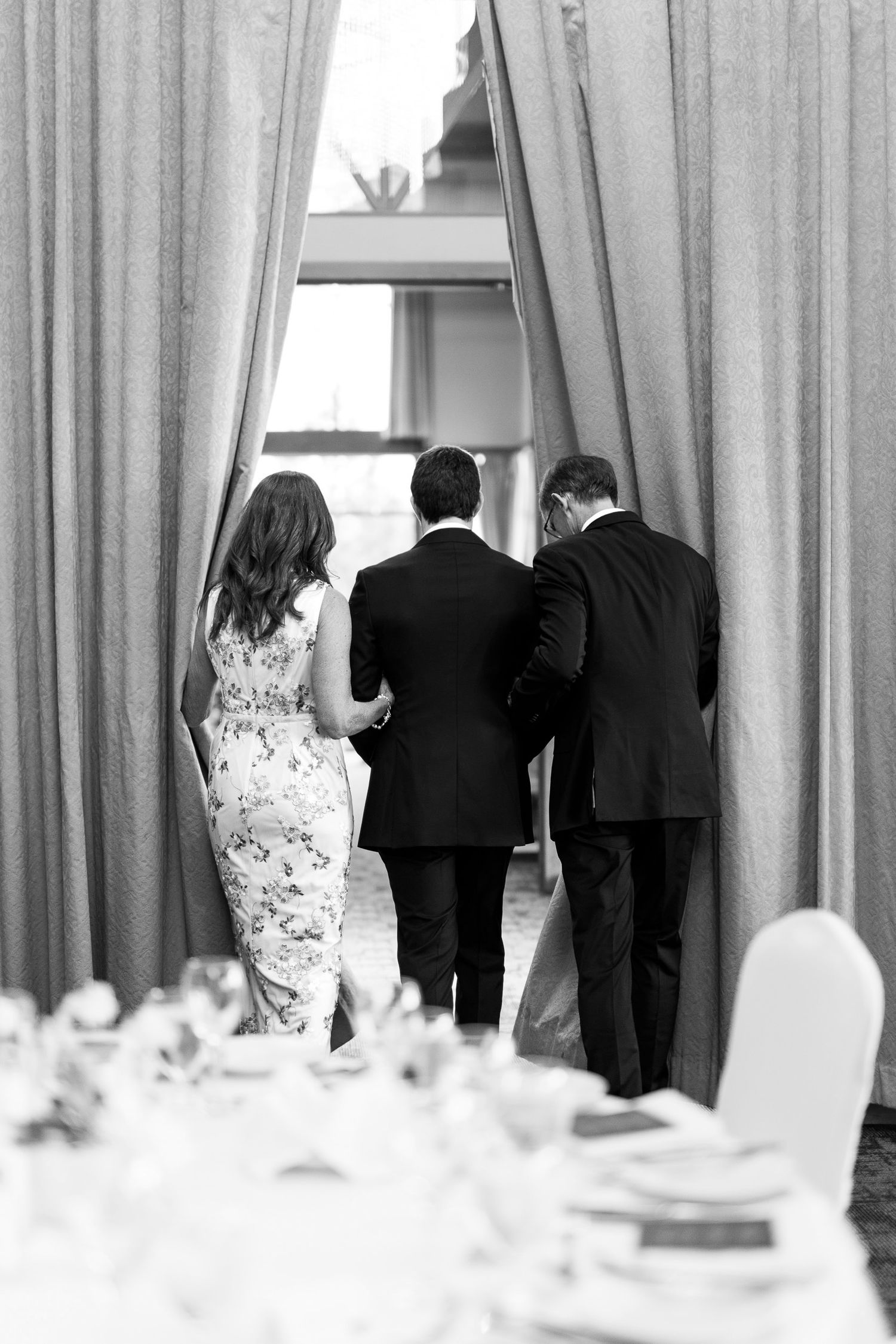 Black and white photo of groom escorted by parents through draped curtains at elegant wedding reception.