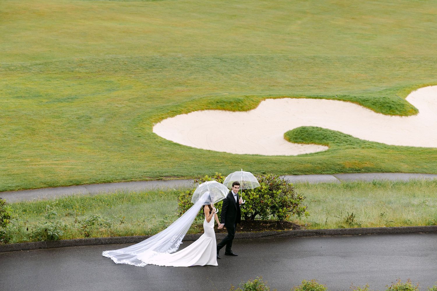 Bride and groom walking on golf course path holding clear umbrellas, wedding veil flowing in wind.