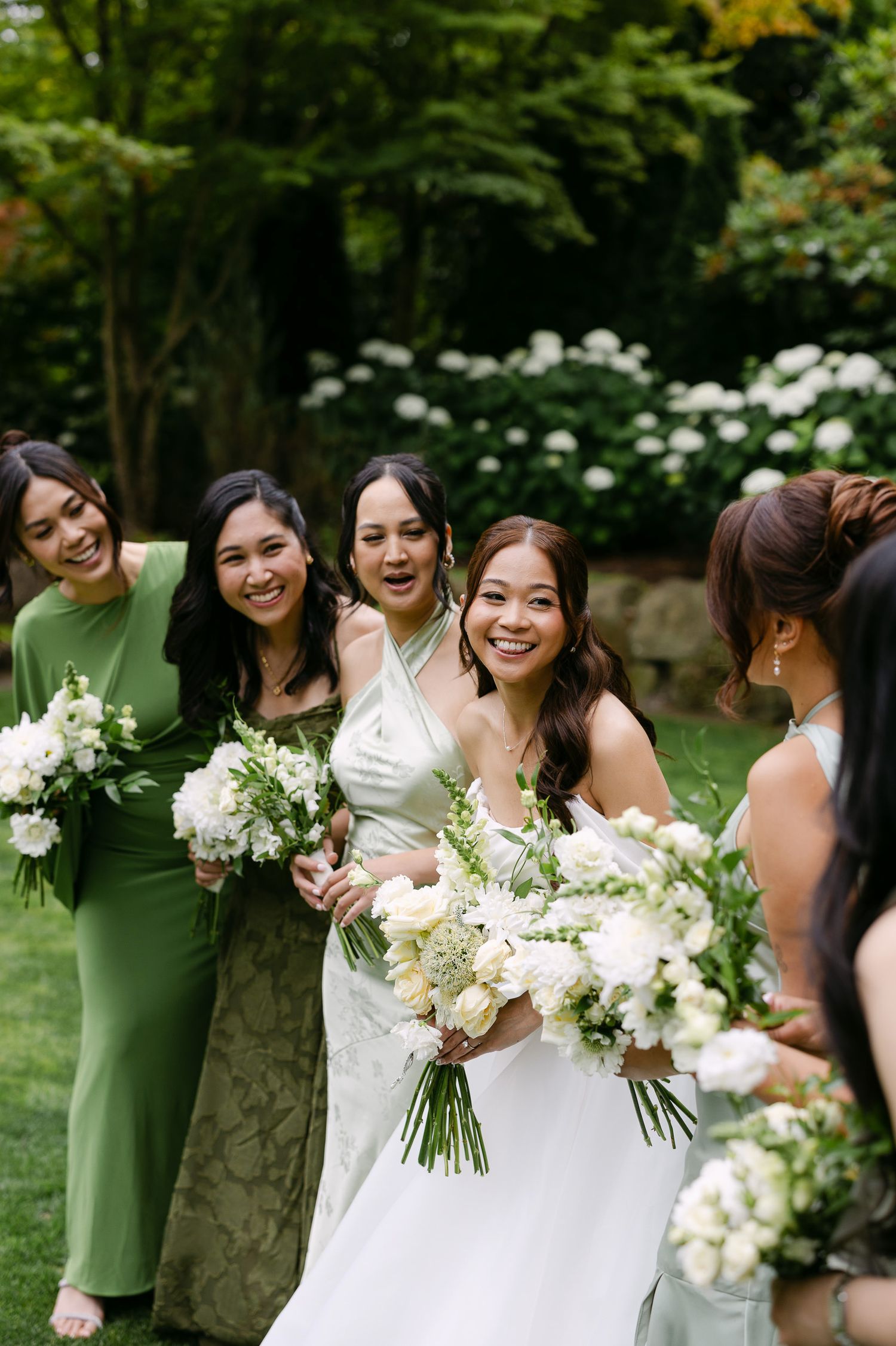 Bride in white gown smiles with bridesmaids in green dresses holding white floral bouquets outdoors.