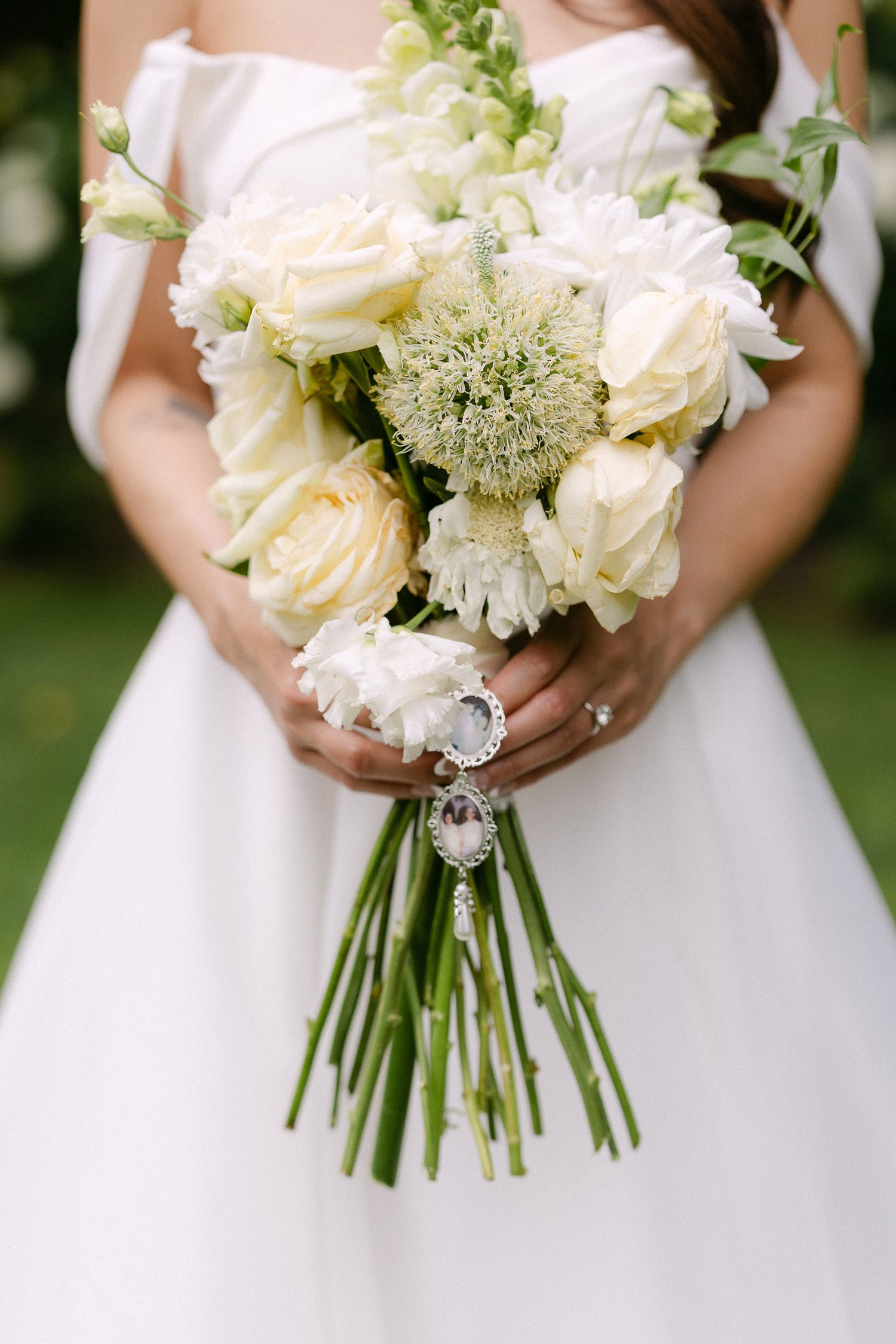 Bride holding a white wedding bouquet with roses and greenery, accented with a silver photo charm pendant.