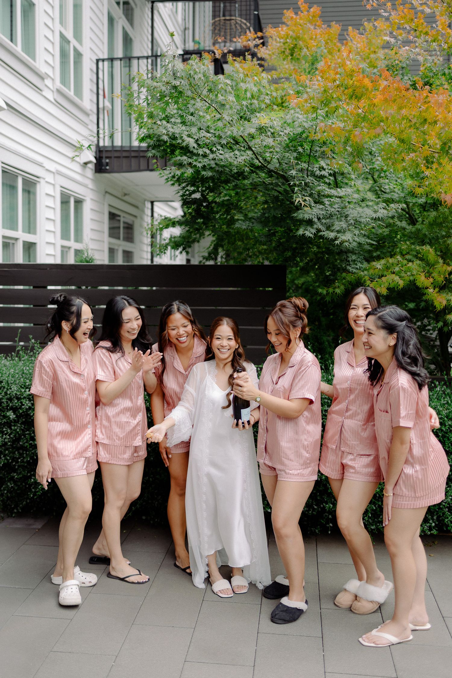 Bride in white robe laughing with bridesmaids in pink satin pajamas while popping champagne outside.