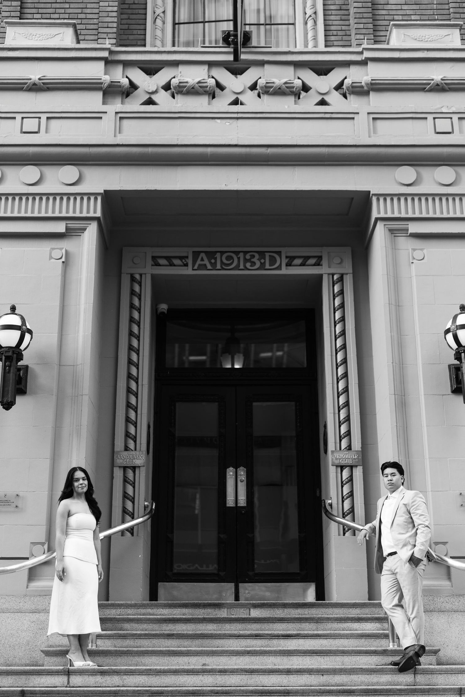 Black and white engagement photo of a couple in formal attire standing on the steps of a historic building with an ornate A-1913-D entrance.