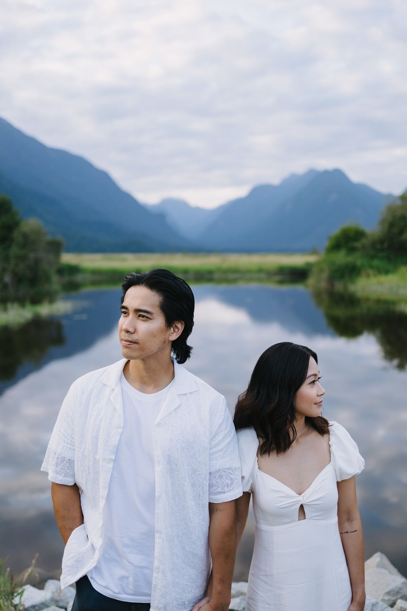 A couple in white outfits stand by a calm reflective lake with misty mountains and cloudy sky behind them.