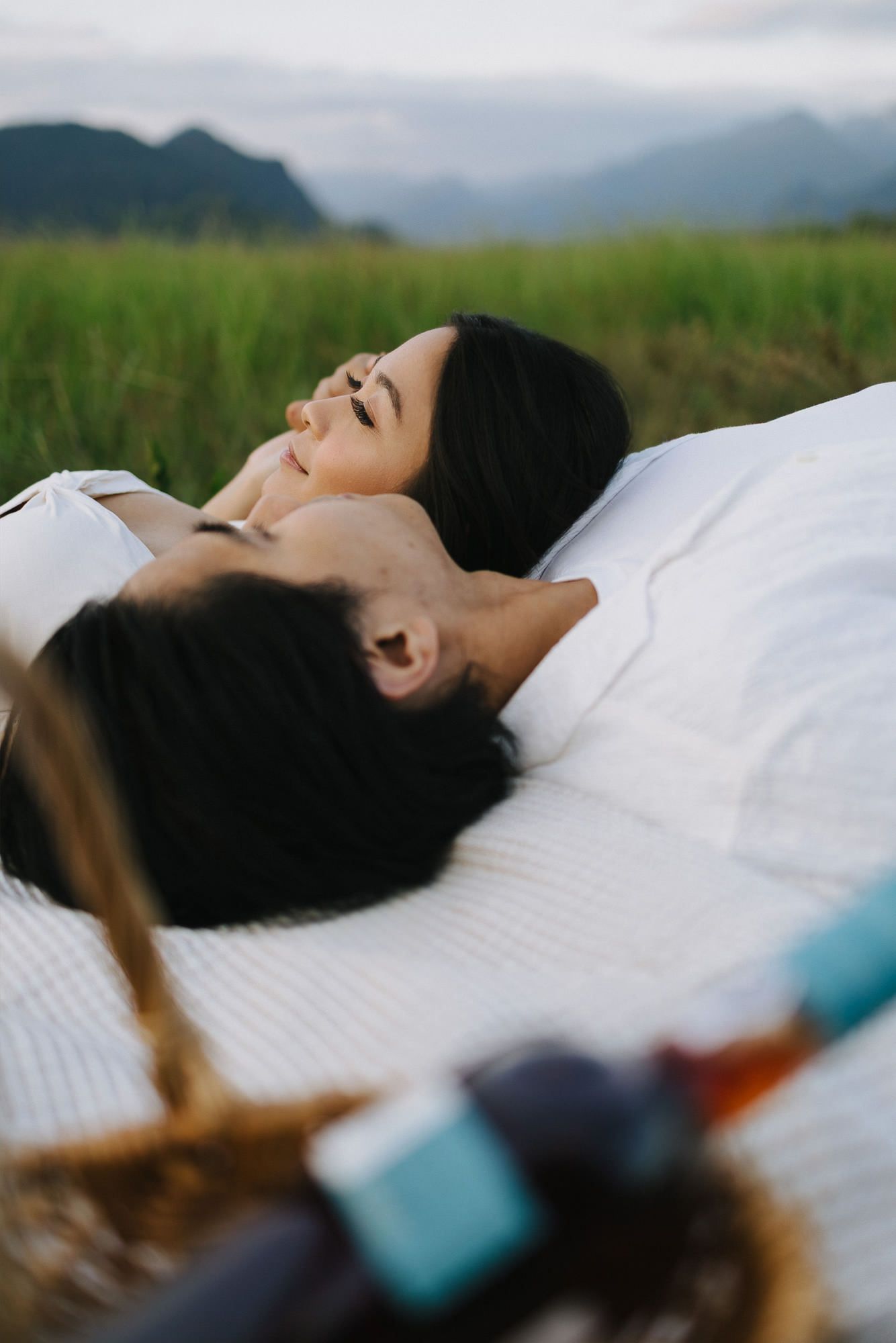 Couple lying close together on a white picnic blanket in a green meadow with mountains in the background and a wicker basket nearby.