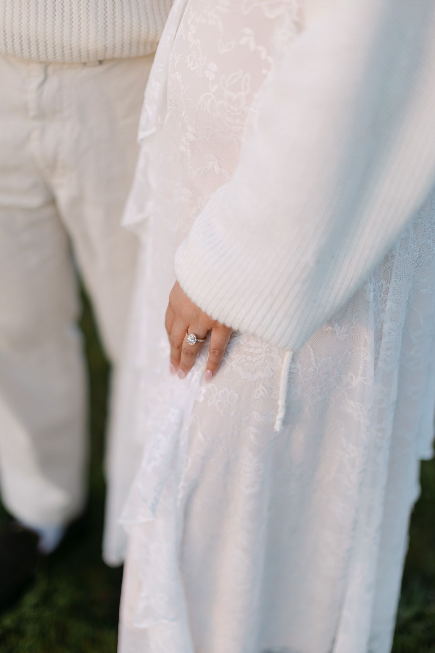 Close-up of a woman's hand with a diamond engagement ring, wearing a white lace dress and cozy knit cardigan beside a person in cream pants.