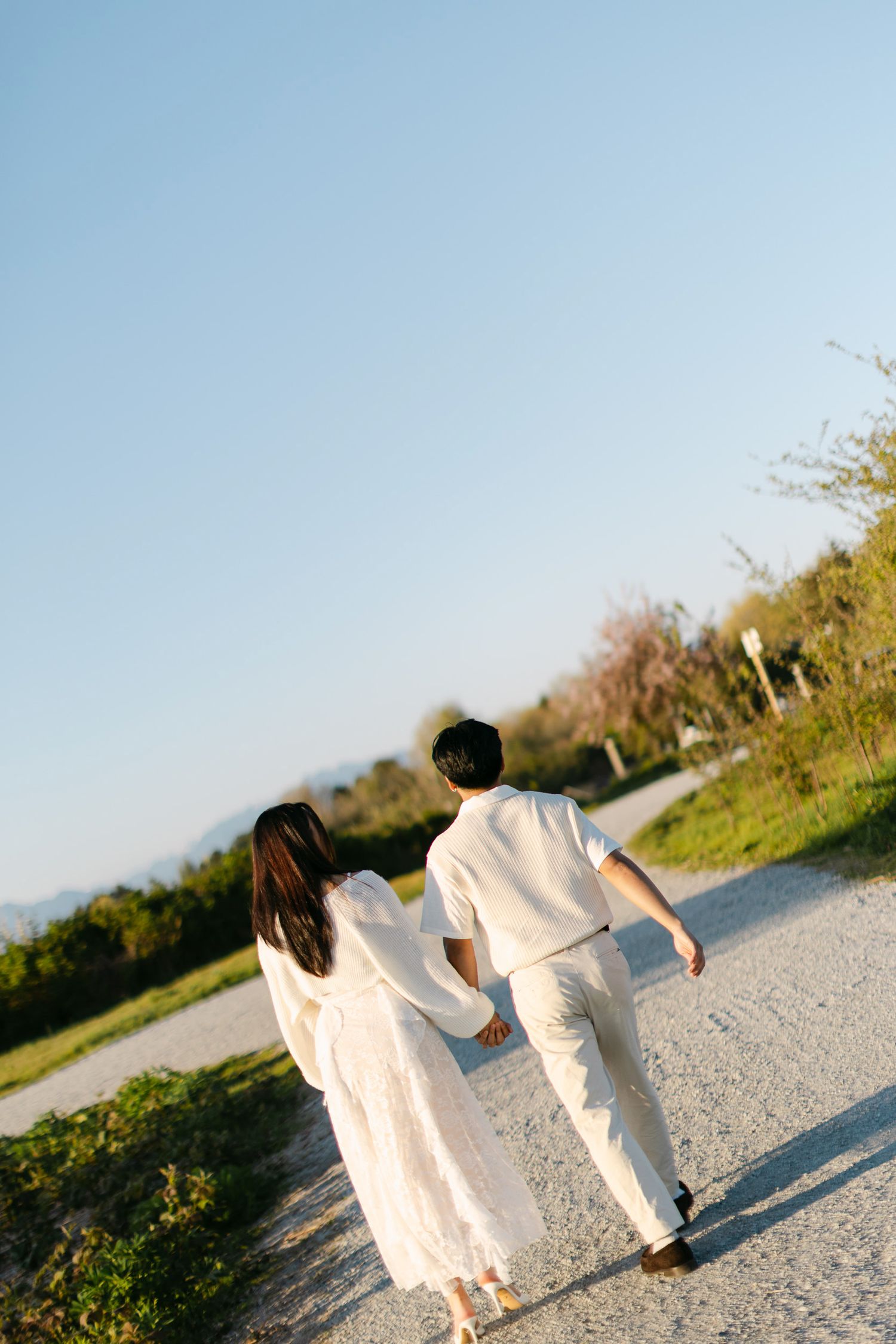 Couple in white clothing walking hand in hand away from the camera down a gravel path lined with trees under a clear blue sky.