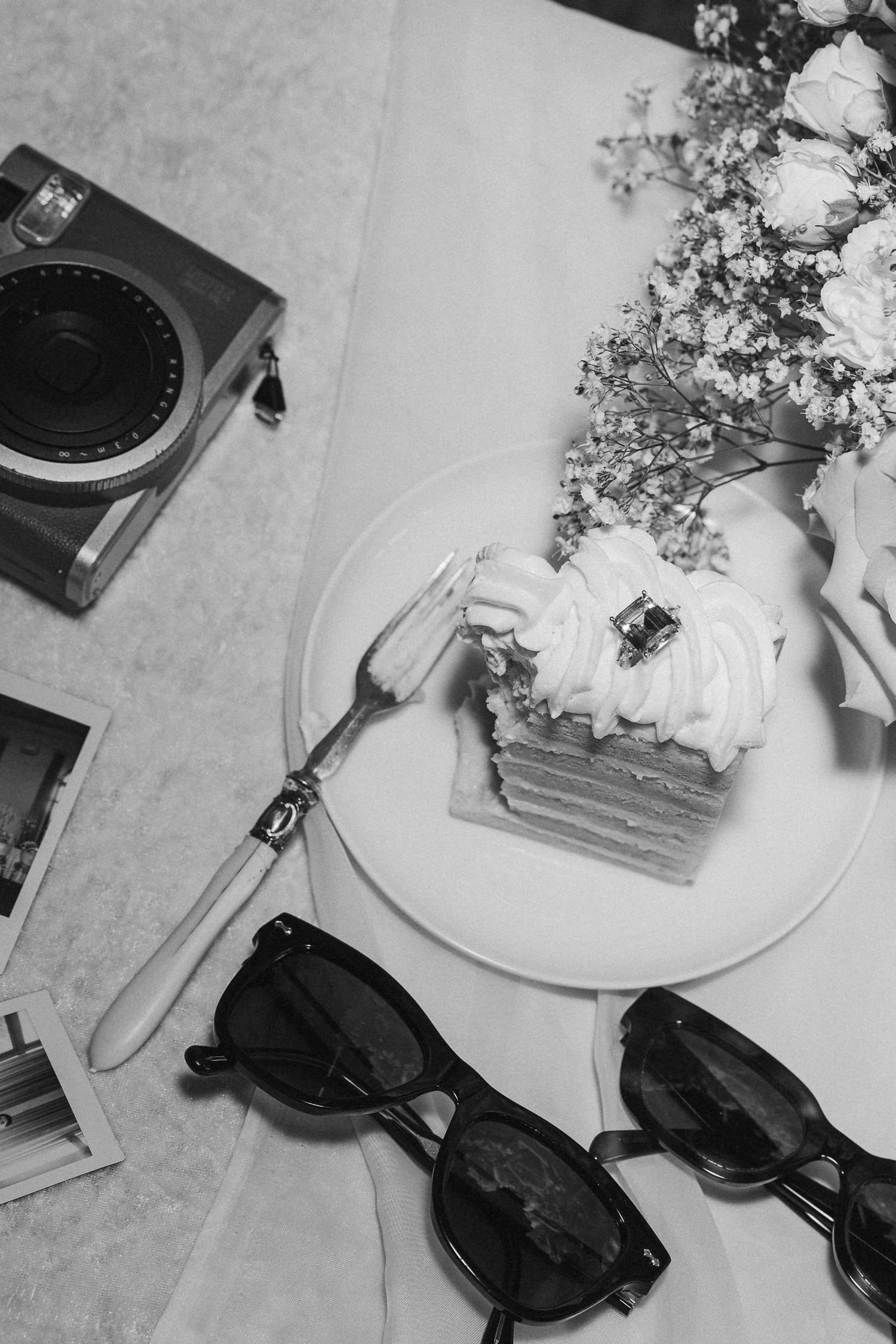 A black and white photo of dessert on a plate with sunglasses and a vintage camera on a table.